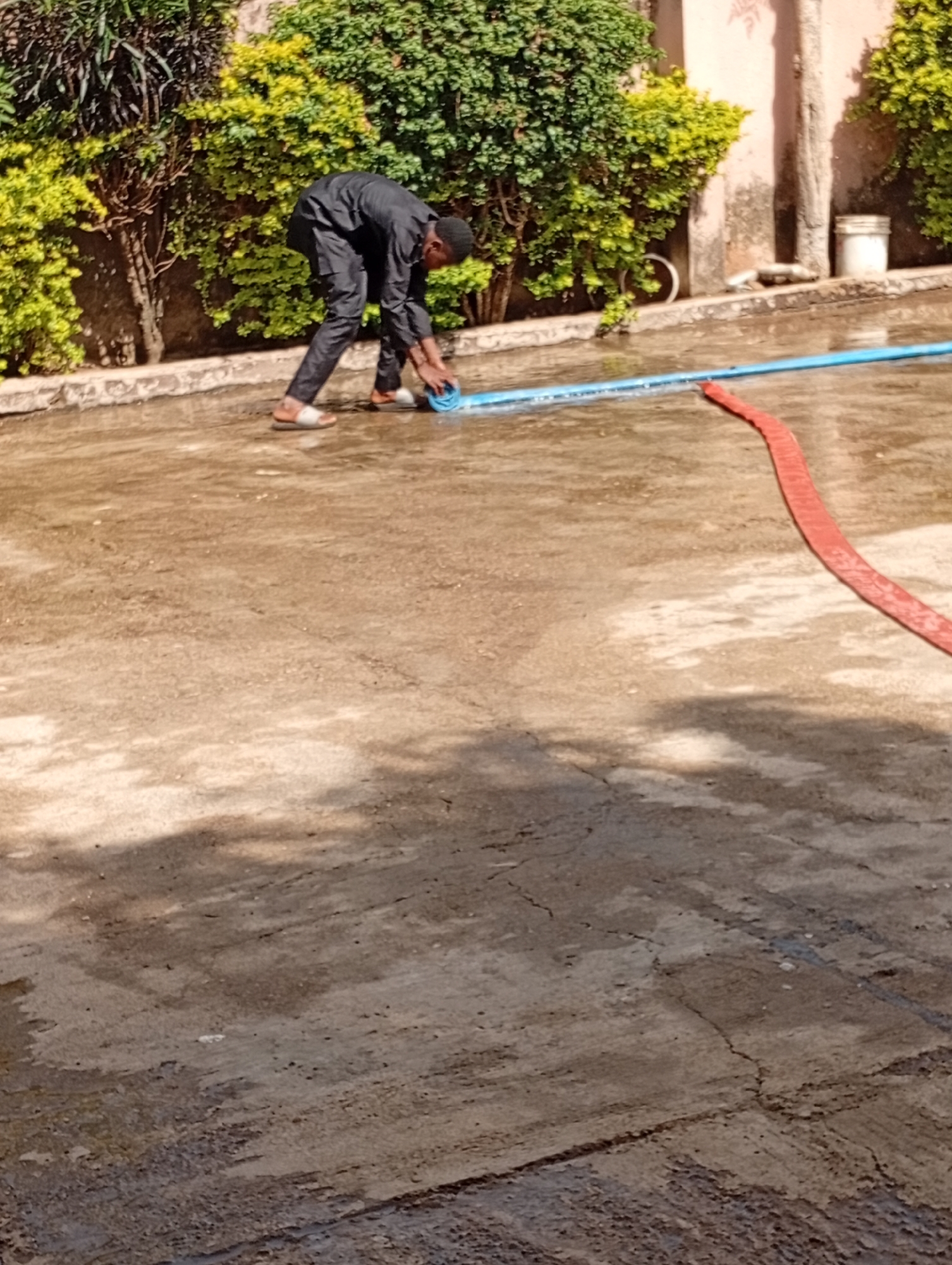 The media file shows a young man dressed in a black outfit, including a long shirt and trousers, and white sandals. He is bending over and appears to be working with a blue hose. Next to him, a red hose is visible on the wet ground. The ground itself is a concrete surface, wet and reflecting the surrounding environment. In the background, there are lush green bushes and trees, suggesting an outdoor setting, possibly a garden or courtyard. A pale pinkish-orange wall and a section of a building are also visible. A white bucket is placed near the wall. The lighting indicates it is daytime, with shadows suggesting the sun is present. The overall scene appears to be a somewhat mundane task, perhaps cleaning or watering. There is no visible text in the media file. The location context is Rafin Par, Nigeria.
