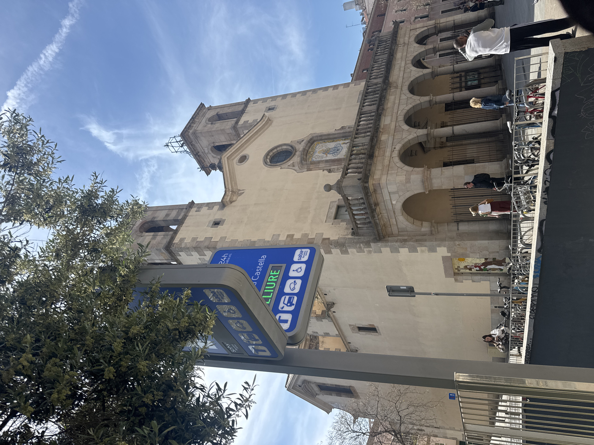 The image captures an upward-angled view of a vibrant street scene in Barcelona, Spain, under a bright blue sky with wispy clouds. The central feature is a grand, light-colored historical building, likely a church or a significant public edifice, with a prominent tower on its left. The facade is richly detailed with numerous arched windows, decorative stone balustrades, and a striking mosaic art piece depicting a scene with figures and a sun, positioned above a distinctive round window.
In the mid-ground, a modern blue digital sign is partially obscured by the lush green foliage of a tree on the left, which bears small pale flowers. The sign, which appears to be for a parking facility, prominently displays "LLIURE" (meaning "available" in Catalan) in large green letters. Other visible text includes "24h", "saba.es", "Castella" (partially visible), the "saba Bamsa" logo, and various icons representing accessible parking, cars, motorcycles, and buses. A numerical display such as "4:35" is also visible.
Below the main building, at street level, several people are present. A person in a white top and dark pants stands near a row of metal bicycle racks. Another individual with blonde hair in a denim jacket is also by the racks. Further down the street, a person in a dark jacket is seen, and a woman with long dark hair, dressed in a white top and a patterned skirt, sits or leans by a metal railing. Numerous bicycles are parked in racks along the street. The bright daylight indicates it is likely morning or afternoon. The architectural style and the visible Catalan text on the sign firmly establish the urban European setting.