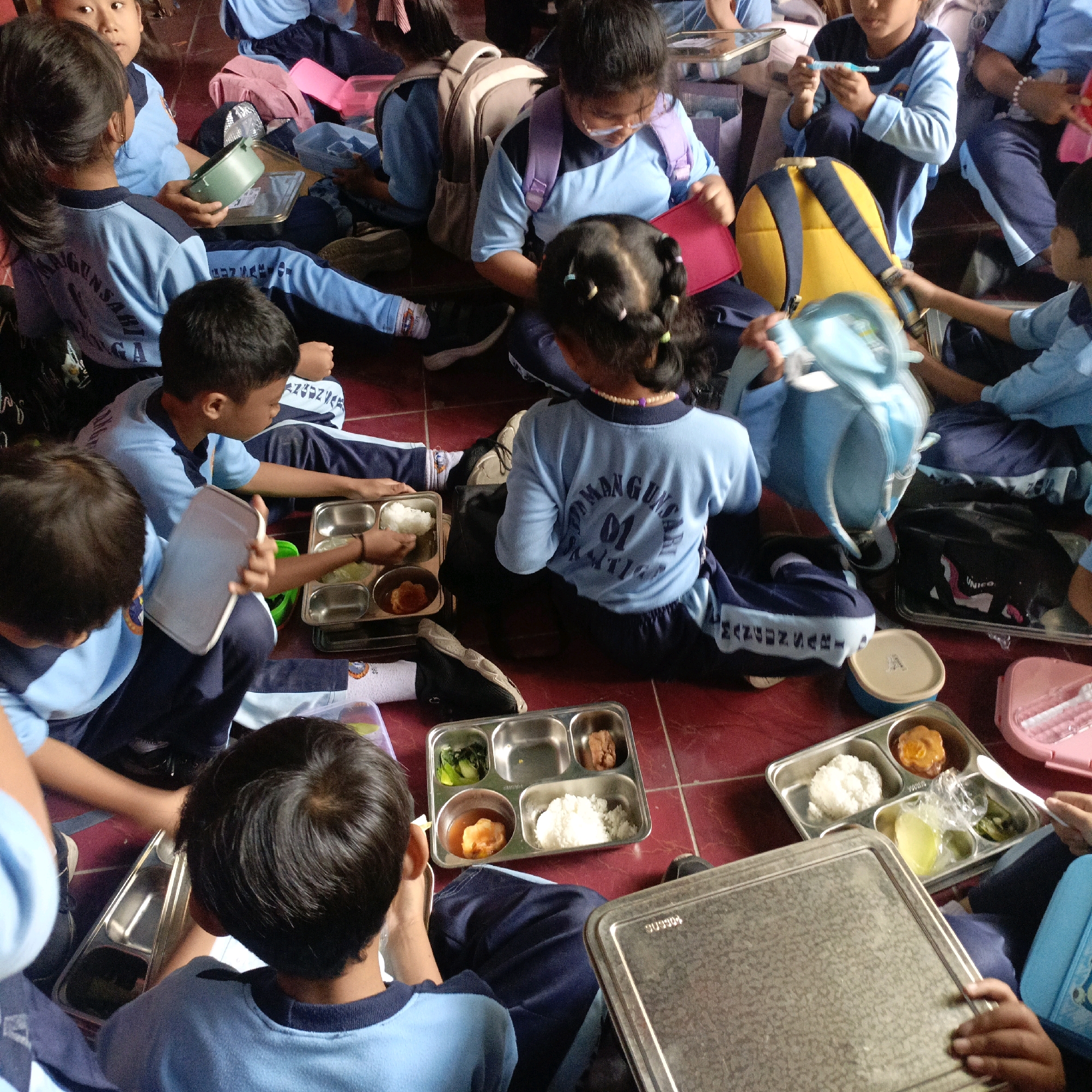 The image depicts a group of young students, likely in a school setting in Salatiga, Indonesia, seated on a red tiled floor, engaged in eating a meal. The students are dressed in light blue shirts and dark blue pants, some with navy blue accents. They are using metal lunch trays with multiple compartments containing rice, vegetables, and what appears to be a piece of meat or fried food. Some students are holding their trays, while others have them on the floor in front of them. Various objects are scattered around, including backpacks, lunchboxes, and a pink plastic container. The atmosphere appears to be casual and communal, with the students focused on their food. The lighting suggests it is daytime, possibly indoors given the lack of strong shadows. The presence of Indonesian writing on some of the students' uniforms confirms the location.