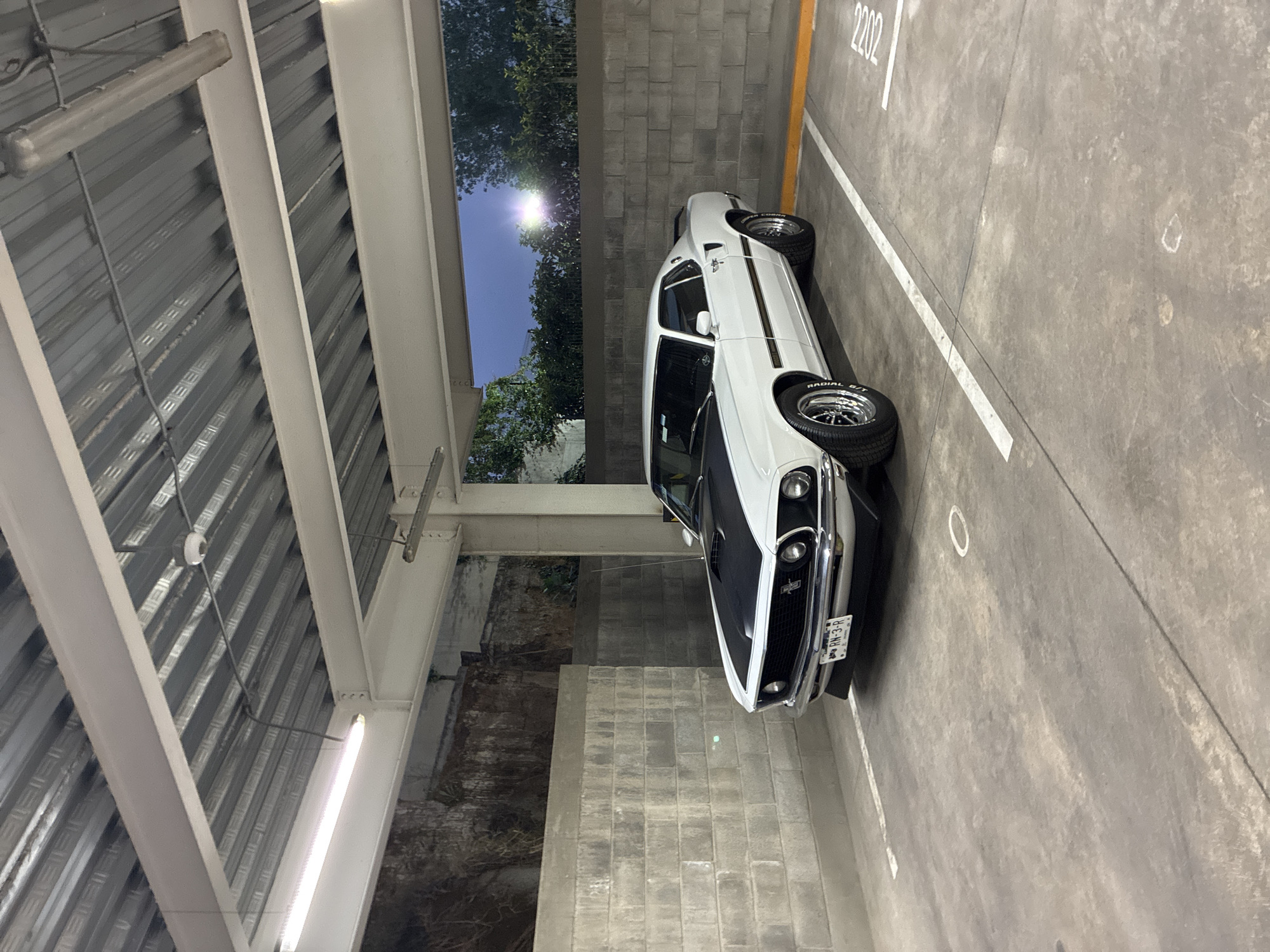 The image shows a white, vintage Ford Mustang parked in parking space 2202 within an indoor parking structure. The structure has a high, corrugated metal ceiling supported by white steel beams and bright fluorescent lights. The surrounding walls are constructed of grey concrete blocks. Outside the parking structure, trees and the sky are visible. The license plate on the car reads "RH-3-R." The location is determined as Huixquilucan de Degollado, Mexico, based on available geolocation data.