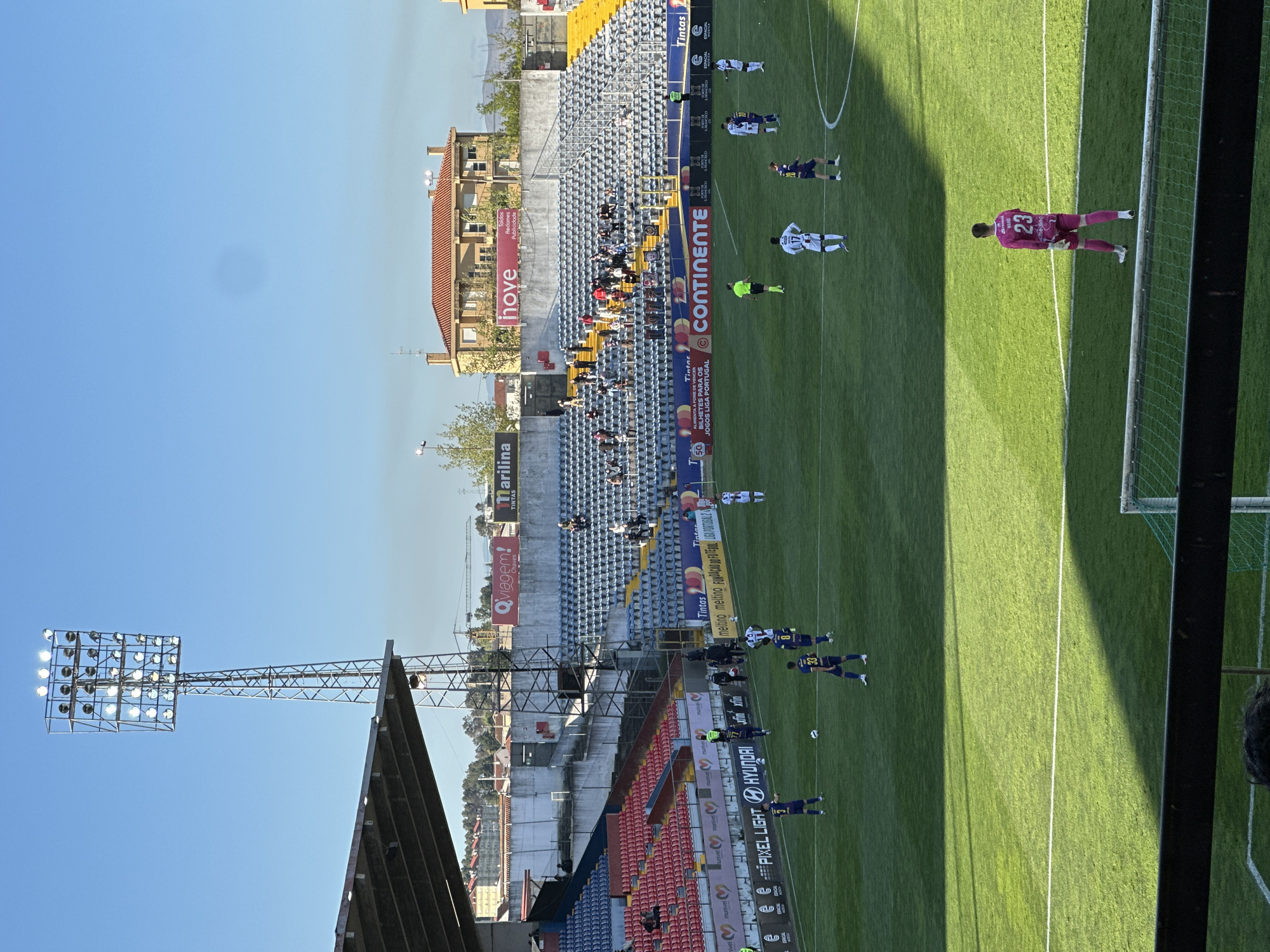 The image shows a soccer match at Estádio Municipal Engenheiro Manuel Branco Teixeira, located in Chaves, Portugal. Players in white and navy uniforms are on the green field, along with a goalkeeper in a pink jersey with the number 23. On the sidelines, branding and advertisement banners are visible. The stadium seating consists of blue and red sections, with a few people in the stands. Behind the stadium, buildings and trees are present. The weather is sunny, and a large stadium light fixture stands tall on the left.