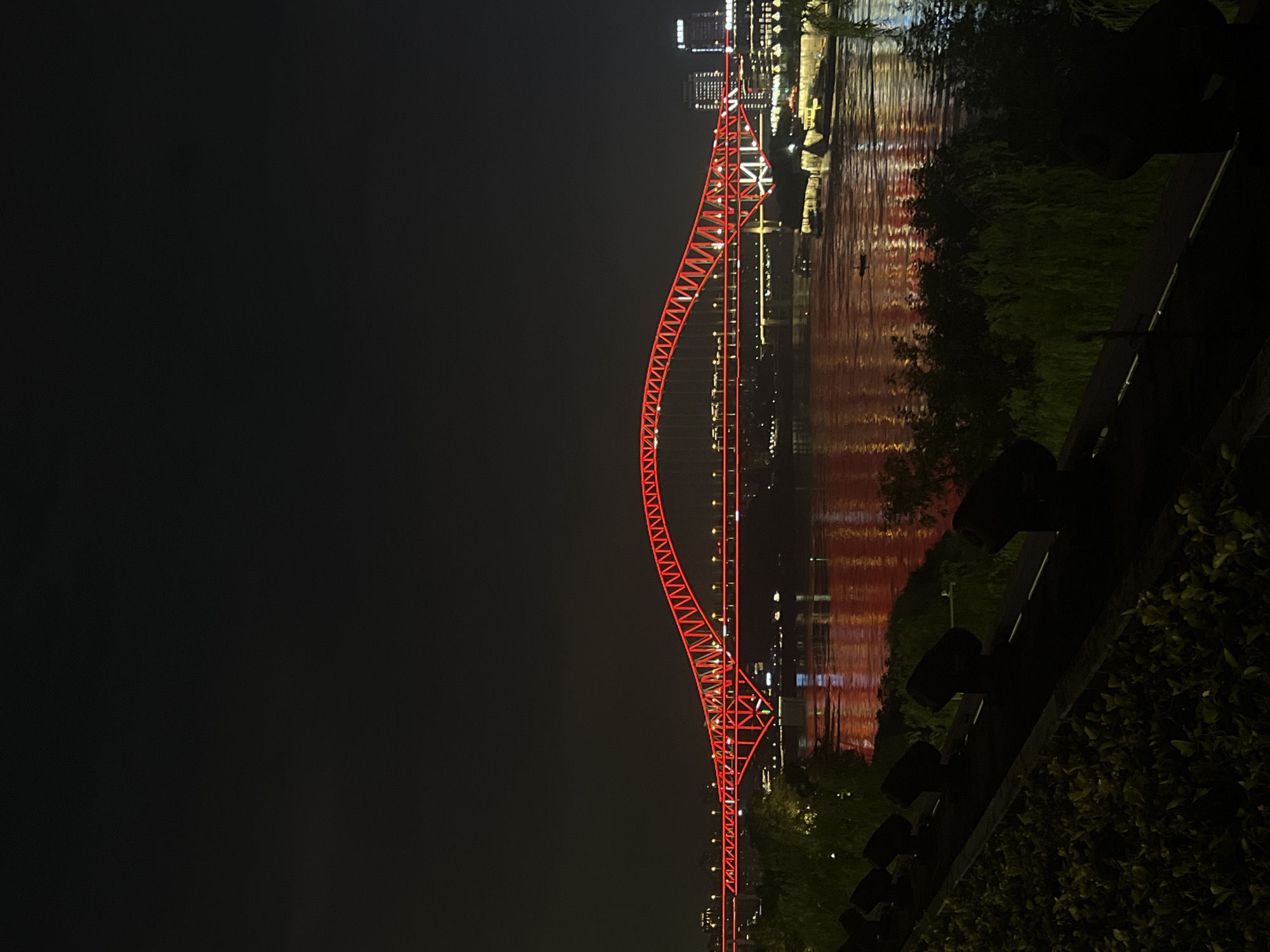 The image depicts a nighttime view of the Yangtze River in Jiangbeicheng Subdistrict, China, featuring a vibrantly lit red arch bridge. The bridge is the central element, illuminated with bright red lights that cast a reflection on the dark, rippling water below. Along the riverbanks, various buildings and lights are visible, indicating an urban setting. In the foreground, a dark, terraced area is lined with black spotlights, and lush green foliage, suggesting a park or viewpoint overlooking the river. The sky is dark and cloudless, consistent with a nighttime scene. The overall impression is one of an illuminated urban landscape at night.