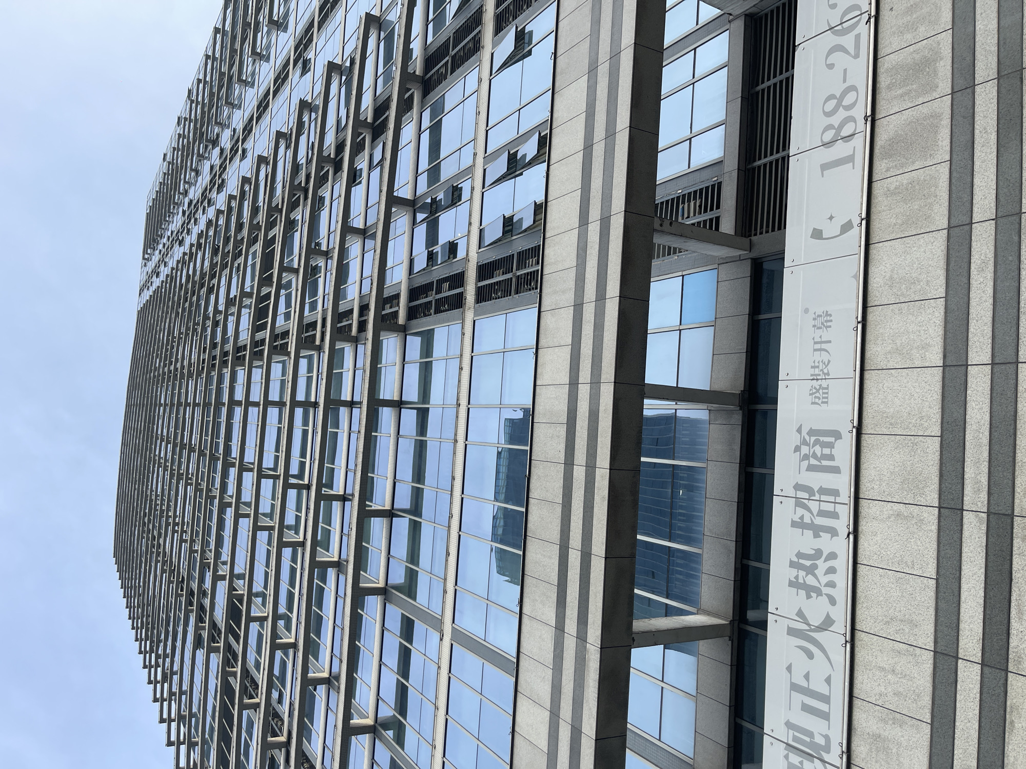 An upward-angled, eye-level shot captures a modern, glass-faced skyscraper in Panyu District, China. The building's facade is a grid of reflective blue glass panes separated by horizontal grey structures that extend outward, creating a layered effect. The sky above is a clear, pale blue, with no clouds visible. In the lower portion of the image, a white banner with grey text is affixed to the building. The text, in Chinese characters, appears to be an advertisement or announcement, with some characters and a phone number visible. Reflections of other buildings are visible in the glass panes of the skyscraper. The image is devoid of any people.