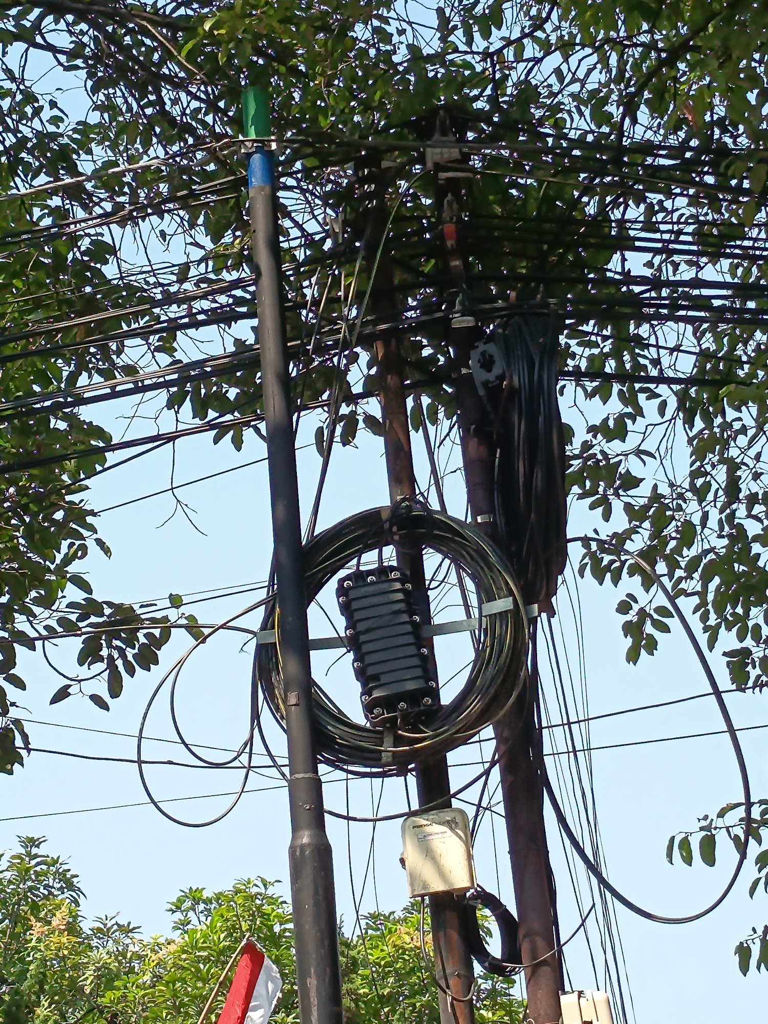 A black utility pole, partially obscured by trees and foliage, stands against a clear blue sky. Multiple electrical wires crisscross the frame, with a bundle of black cables coiled around a gray metal bracket attached to the pole. A black rectangular junction box with silver clasps is situated within the coiled cables. Another smaller, off-white rectangular box with a transparent cover is mounted lower on the pole. A thin, black pipe, capped with blue and green, extends vertically from the top of the pole. In the lower left corner, a small Indonesian flag, red and white, is visible amongst green trees. The scene appears to be in an outdoor, possibly urban or semi-urban, environment in Kandangjati Kulon, Indonesia, given the presence of utility infrastructure and a national flag.