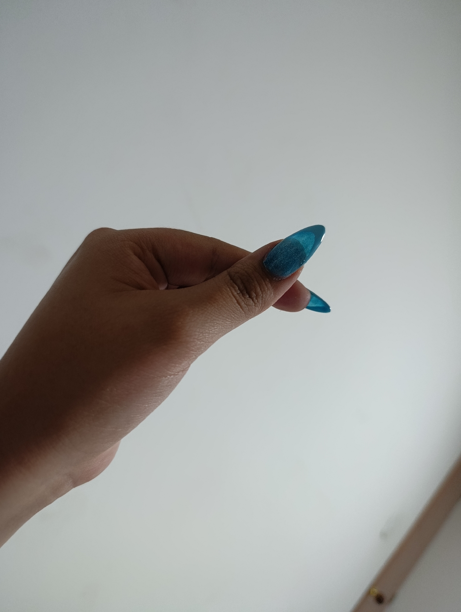 A hand with two prominent teal-colored, stiletto-shaped fingernails is displayed against a blurred off-white background. The hand appears to be of a person with medium-toned skin. The scene is possibly indoors, suggested by the controlled lighting and the indistinct object visible in the lower right corner, which might be a door frame or molding. There is no indication of Funza, Colombia.