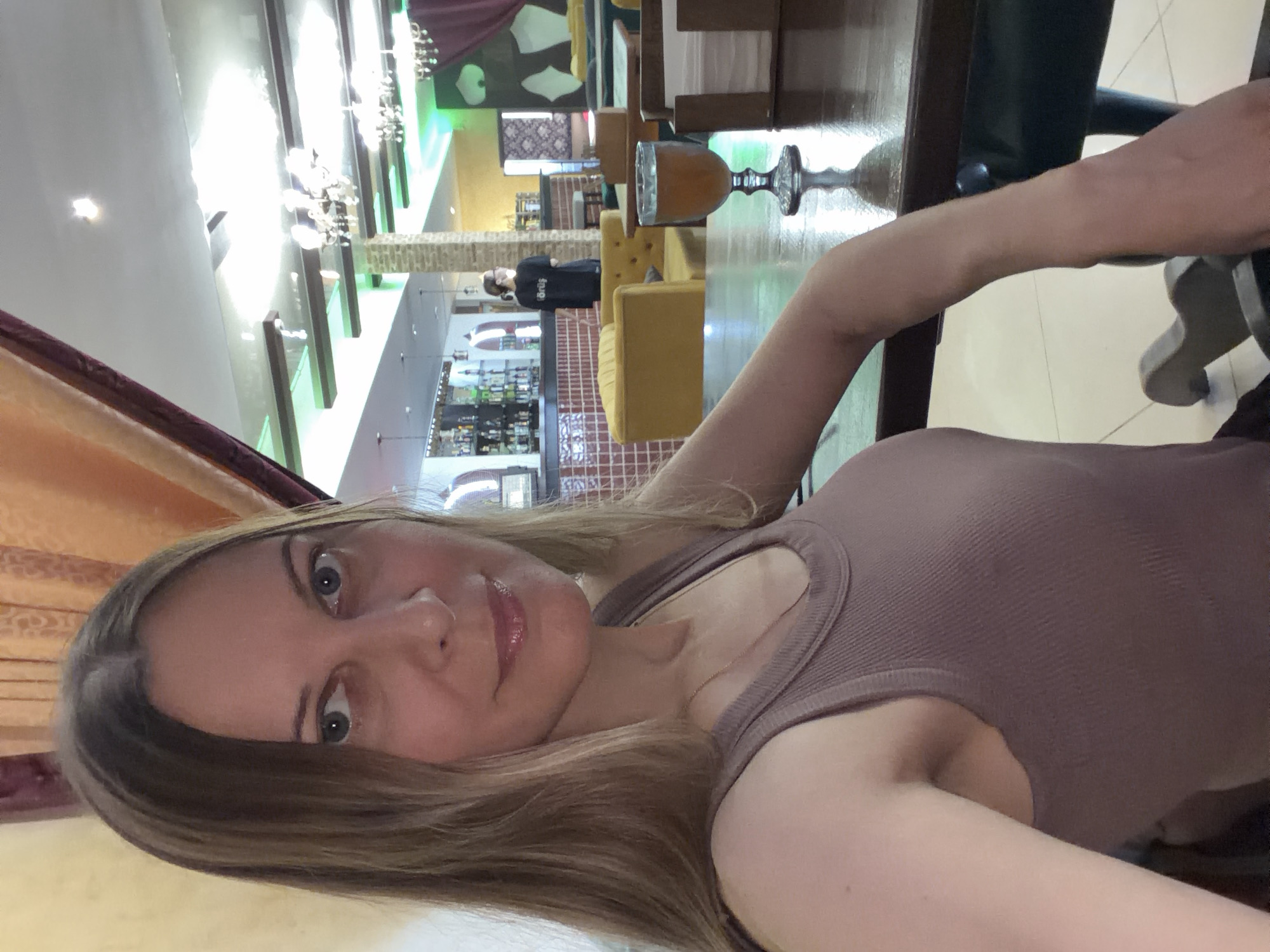 A woman with blonde hair is taking a selfie in a dimly lit restaurant. She is wearing a mauve tank top. A glass of amber liquid sits on a wooden table in front of her. In the background, a bar is visible, as are several tables and chairs. The restaurant appears to be decorated with curtains and ornate light fixtures.