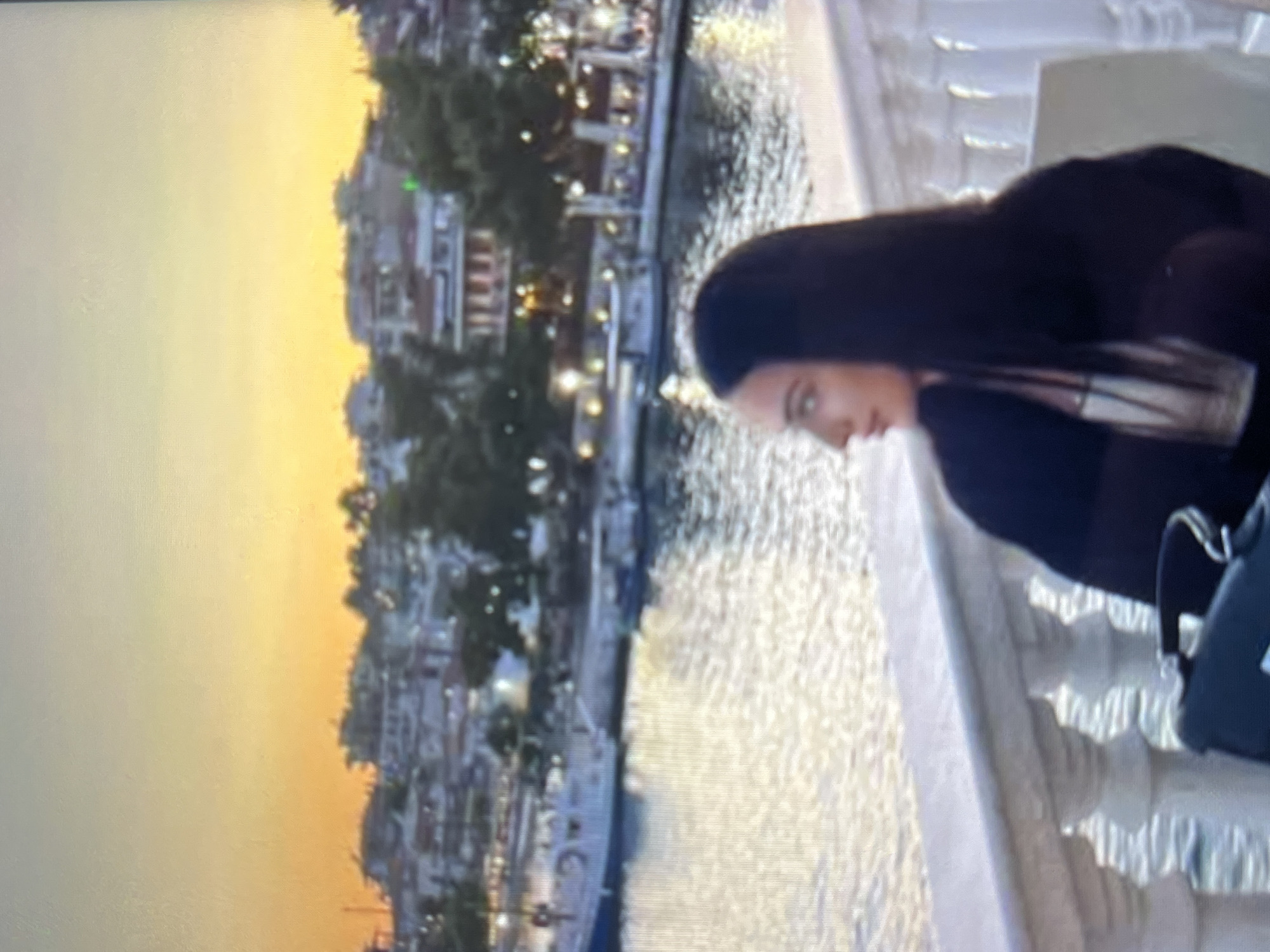 The image captures a woman with long, dark hair in profile, looking towards the left. She is seated at an outdoor venue with a white balustrade in the foreground, suggesting a waterfront or balcony setting. Behind her, a body of water reflects the warm hues of a sunset sky, which transitions from yellow to orange. The background features a cityscape with buildings nestled amongst trees, illuminated by various lights. Boats are visible docked in the water. The overall scene conveys a peaceful, picturesque ambiance during dusk. There is no indication of the location being Vratsa, Bulgaria.