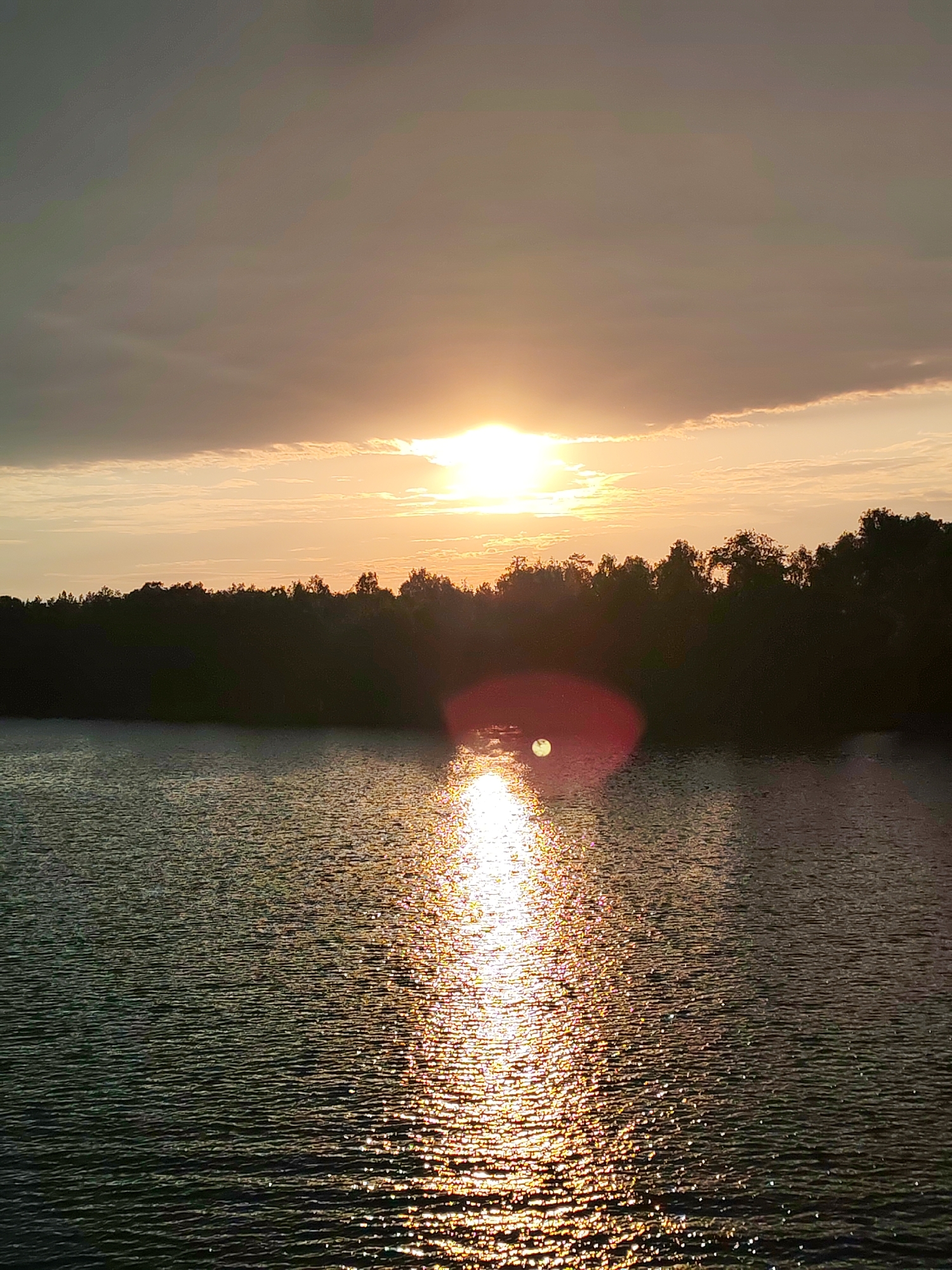 The image shows a sunset over a body of water. The sun is partially obscured by clouds, but its light is reflected in a shimmering path across the water's surface. A dark treeline is visible in the middle ground, silhouetted against the bright sky. No people are visible. The city is not specified.