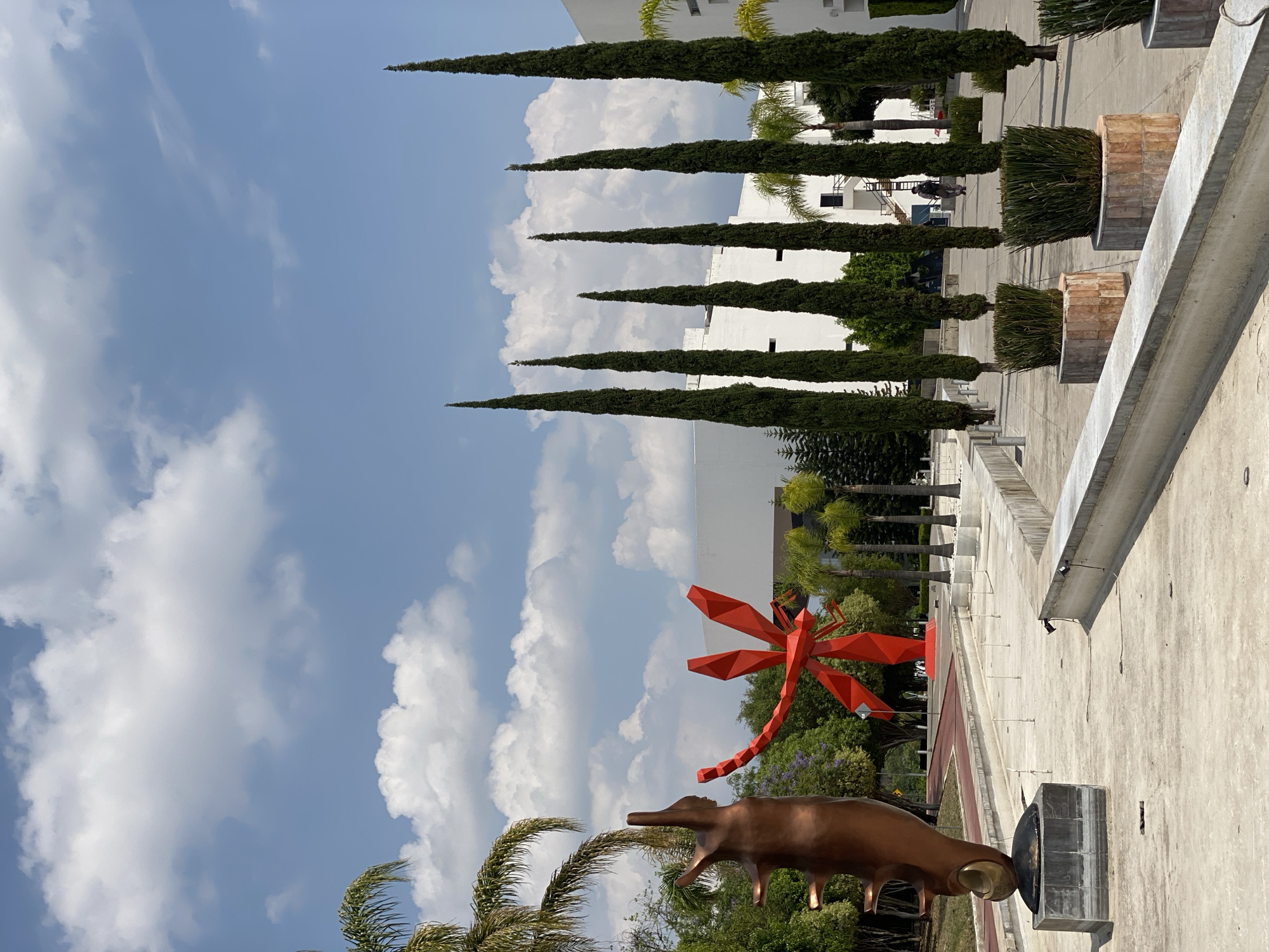 The image depicts an outdoor scene with several notable features. In the foreground, there is a metallic sculpture of a bovine animal with an unusual head, positioned alongside a large, red geometric dragonfly sculpture. Behind these sculptures, lush green trees and foliage can be seen. The sky is a mix of light blue and white clouds.
Further back, a row of tall, slender trees leads the eye towards a white building. There are also large potted plants with square brick designs. The scene is presented from a slightly elevated perspective, capturing architectural details of the wall and surroundings.