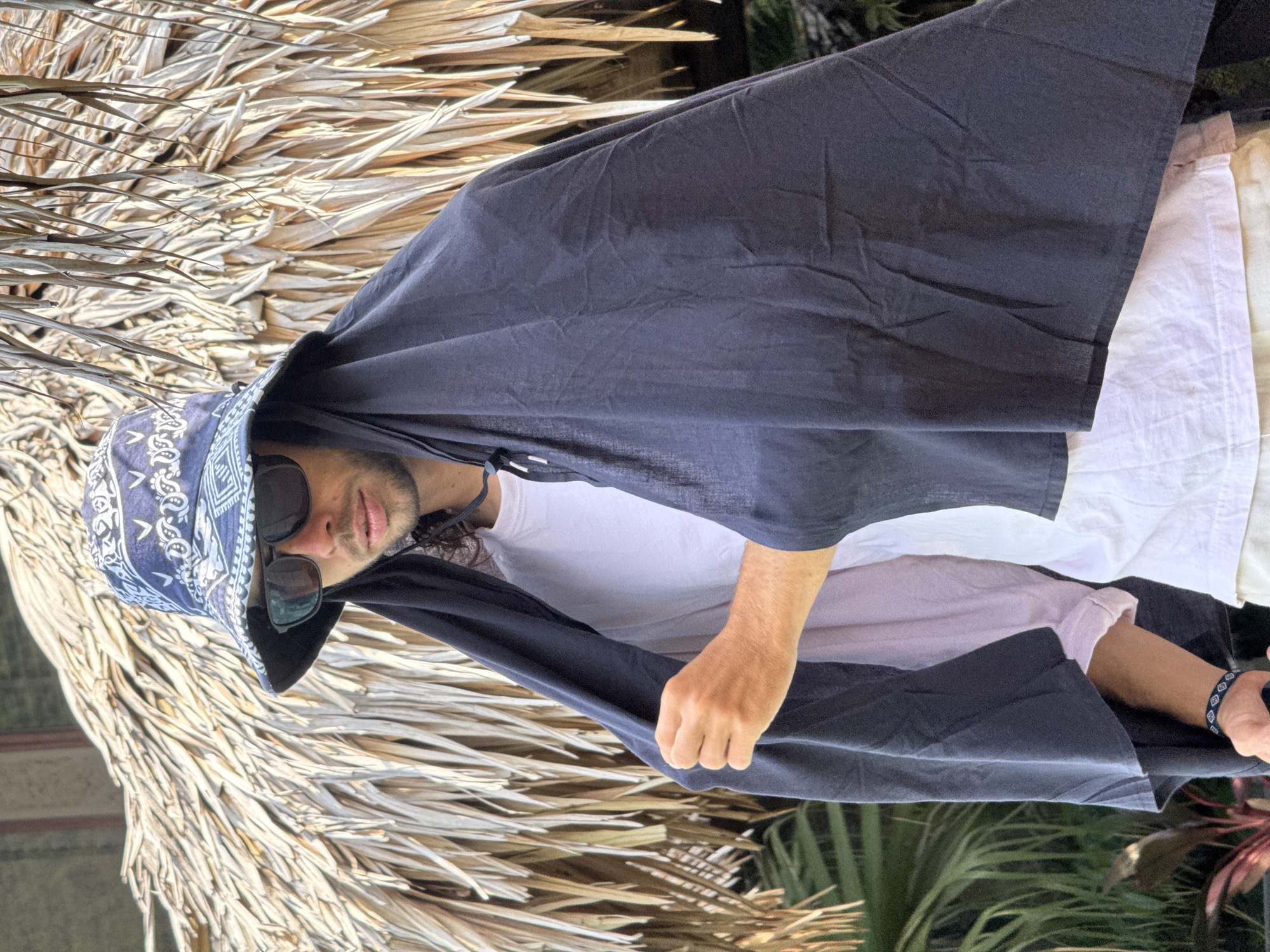 A young man stands outdoors, wearing a blue bandana print bucket hat and sunglasses, with a dark fabric draped over his head and shoulders, resembling a hood. He is wearing a white t-shirt and beige shorts, and has a bracelet on his left wrist. He is holding the dark fabric with his left hand. Behind him is a thatched roof structure, possibly a hut or umbrella, made of dried palm leaves. Lush green foliage and what appear to be palm fronds are visible in the background, suggesting a tropical or sub-tropical environment. The lighting indicates it is daytime, with the sun casting shadows and highlighting the textures of the thatch. The scene is likely a tourist area or a relaxed outdoor setting, possibly in a location with a warm climate. There is no specific indication of this location being in Nam Hoa Lư Ward, Vietnam, but it could be a general representation of a tropical outdoor setting.