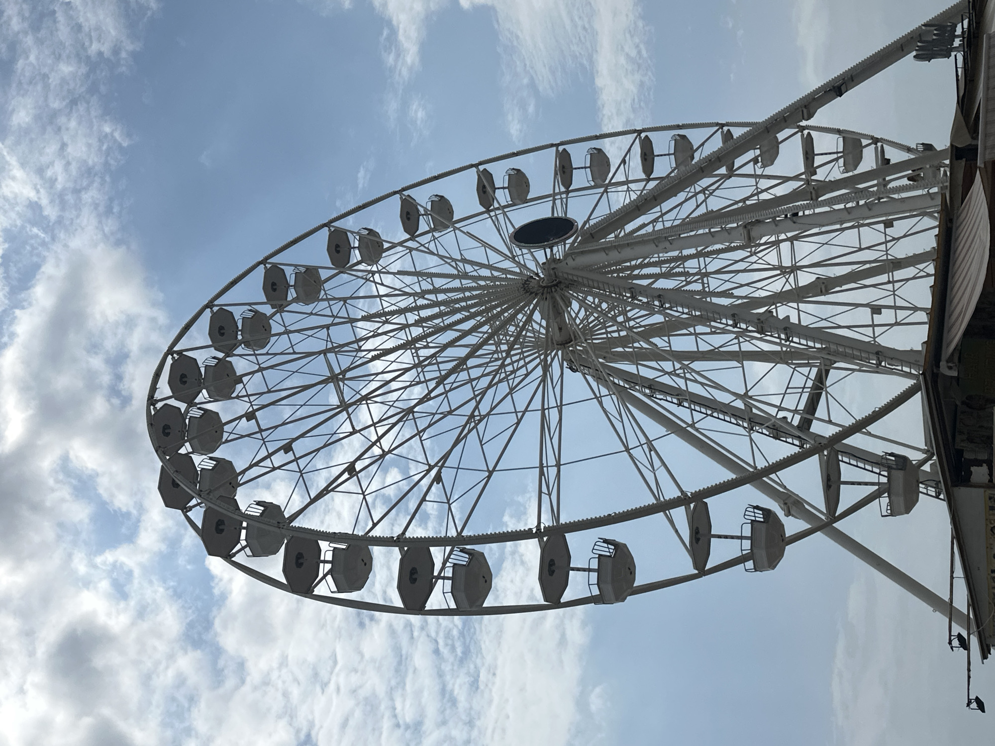 A low-angle shot captures a large, white Ferris wheel against a partly cloudy blue sky. The Ferris wheel's intricate metal structure is visible, with many spokes radiating from the center and numerous gondolas hanging from the outer rim. Some of the gondolas are illuminated. The base of the Ferris wheel rests on a structure that appears to be a market stall or a building with awnings. The scene is set outdoors, likely in a recreational area or amusement park.