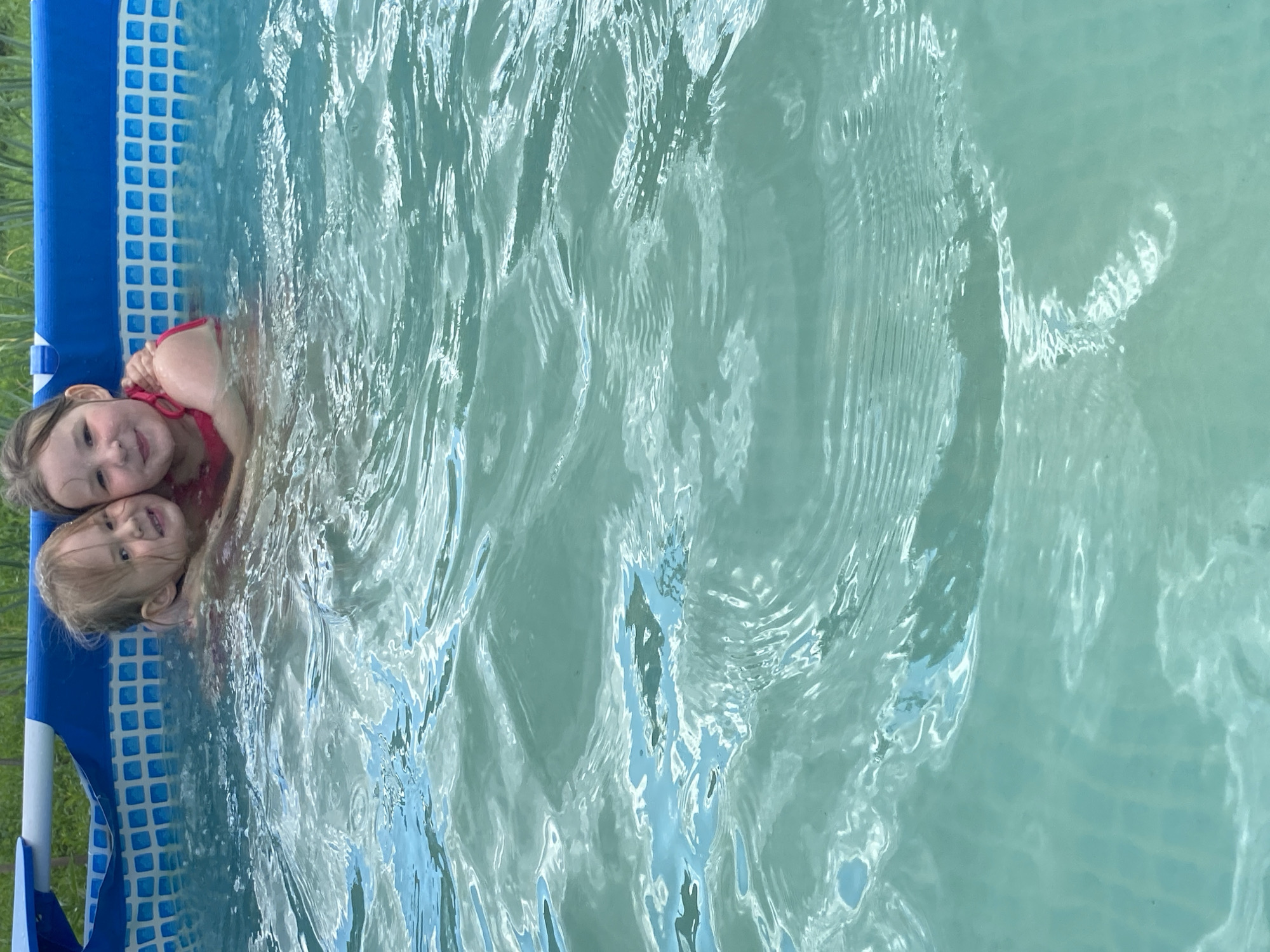 In the city of Chaikovsky, Russia, two young girls are embraced in a bright blue and white tiled above-ground swimming pool. The water is a clear turquoise, with sunlight creating shimmering patterns on its surface. The girls, one with blonde hair and the other with dark hair, are wearing pink swimsuits and smiling at the camera. The pool is surrounded by green foliage, suggesting a backyard setting. The image captures a moment of joy and summer fun.