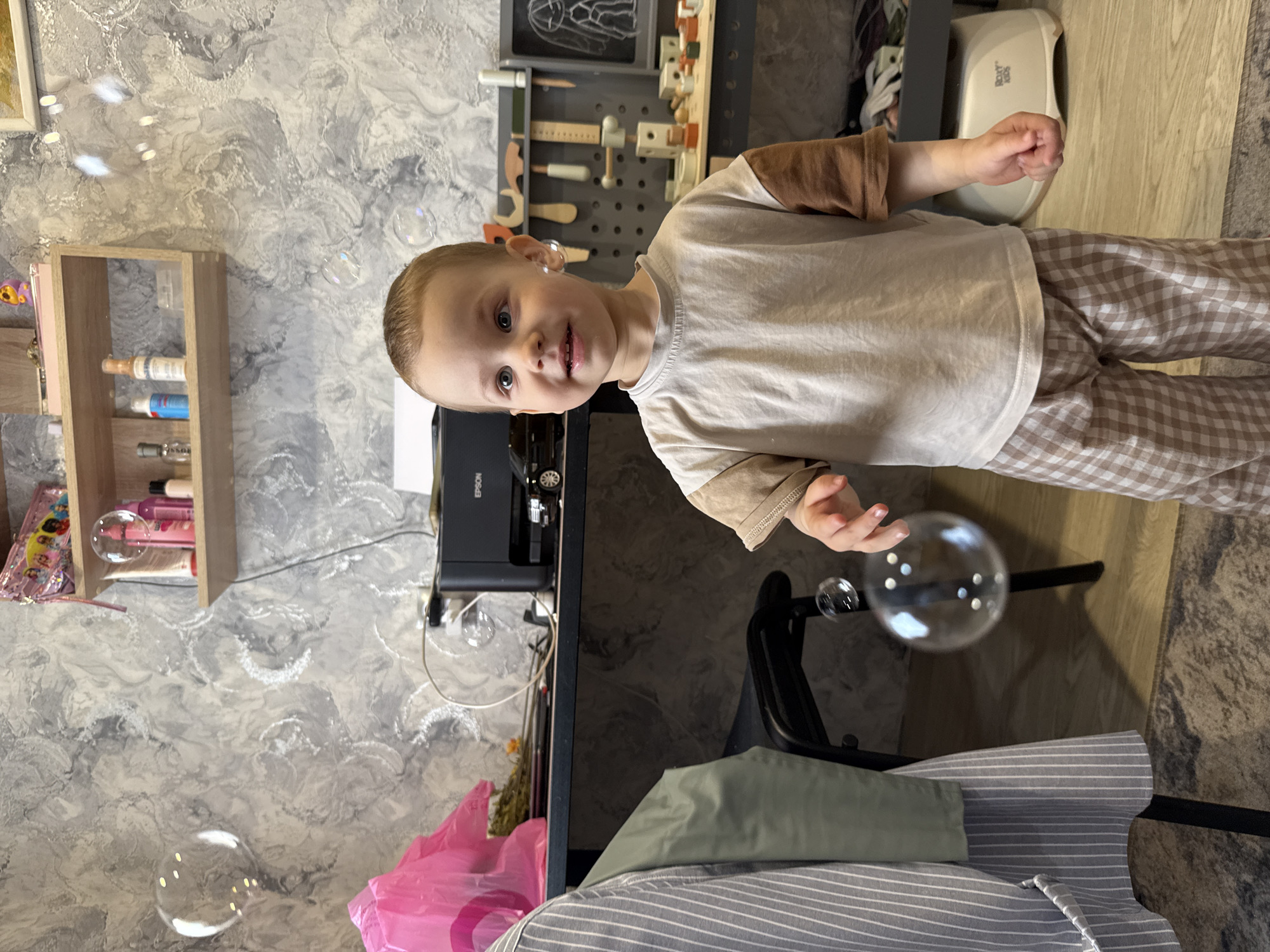 A young child stands indoors, wearing a tan and brown long-sleeved shirt and brown and white checkered pants. The child is partially in front of a desk upon which there is an Epson printer. Behind the desk there is a wall with shelves holding items. There are soap bubbles around the child. There is no clear indication of Rusanovo, Russia in the image.