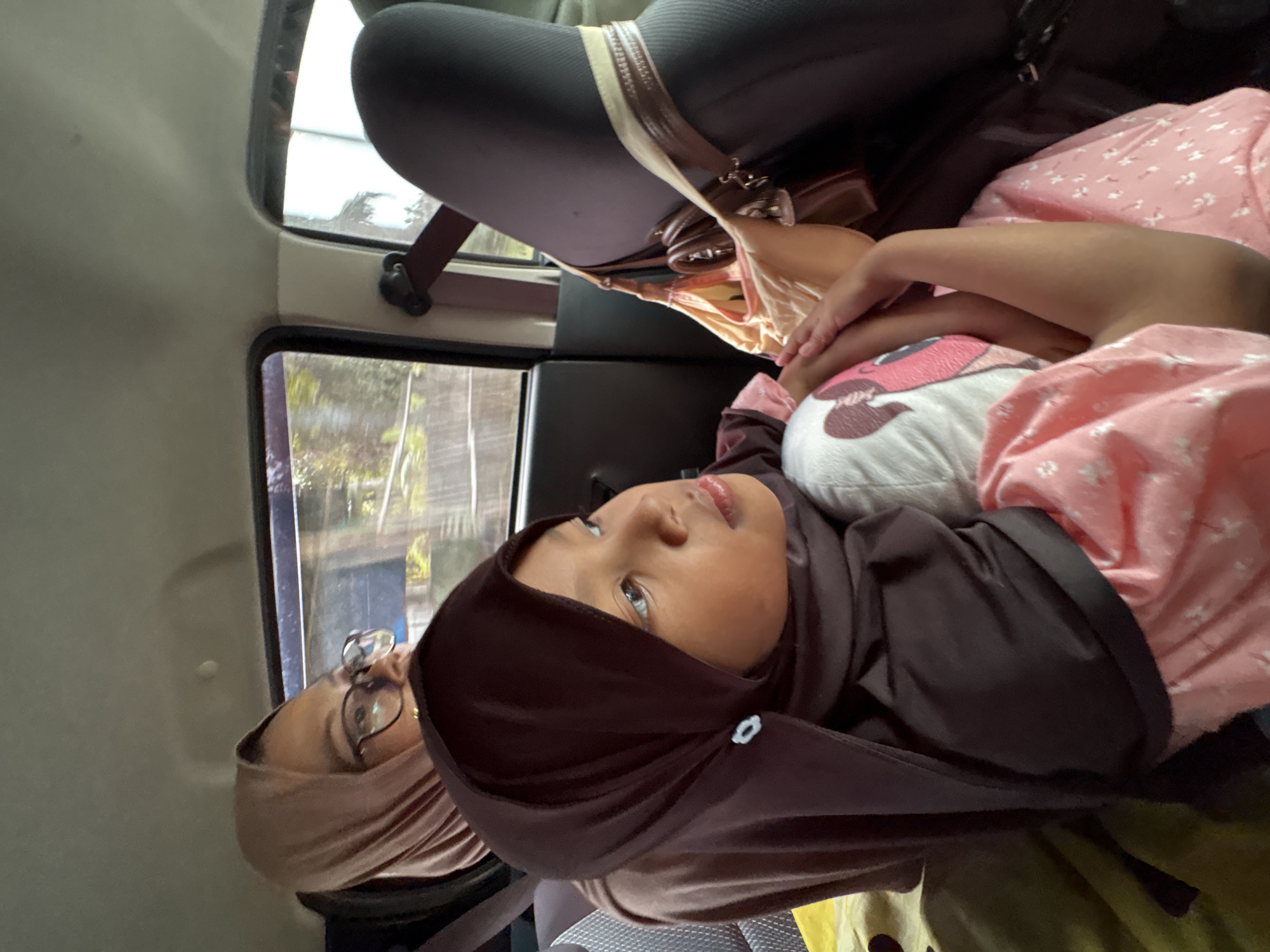 In Gambirono, Indonesia, two individuals are seated in the back of a vehicle. The foreground is occupied by a young girl wearing a dark brown hijab and a pink floral dress. She is holding a plush toy with a cartoon character. Behind her, a woman wearing a light pink hijab and glasses is also visible. The interior of the car, including a seatbelt and a handbag, is also depicted. The view outside the car windows suggests a tree-lined environment, indicating a location with vegetation.