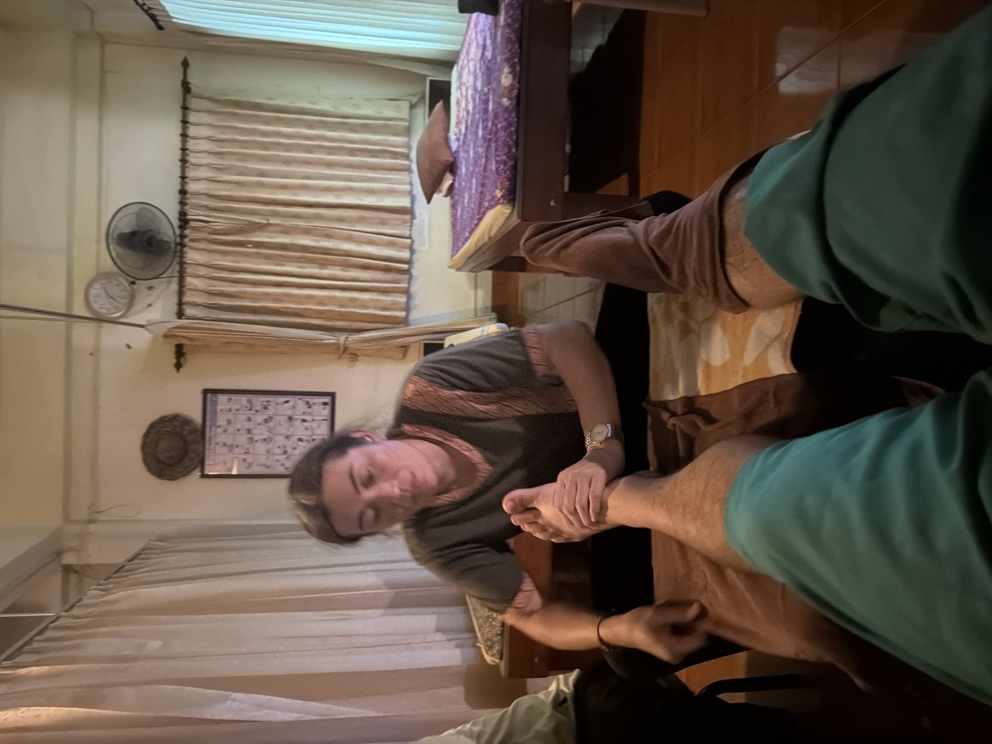 The image depicts a foot massage session in Baan Tai, Thailand. A person, likely a massage therapist, is kneeling and massaging the foot of another person whose legs are visible in the foreground. The therapist is wearing a green shirt with ornate trim. The environment appears to be a massage parlor, with curtains and beds visible in the background. One bed is covered with a purple patterned cloth and a brown pillow. A clock and a fan are mounted on the wall above the curtains. The flooring is wooden.