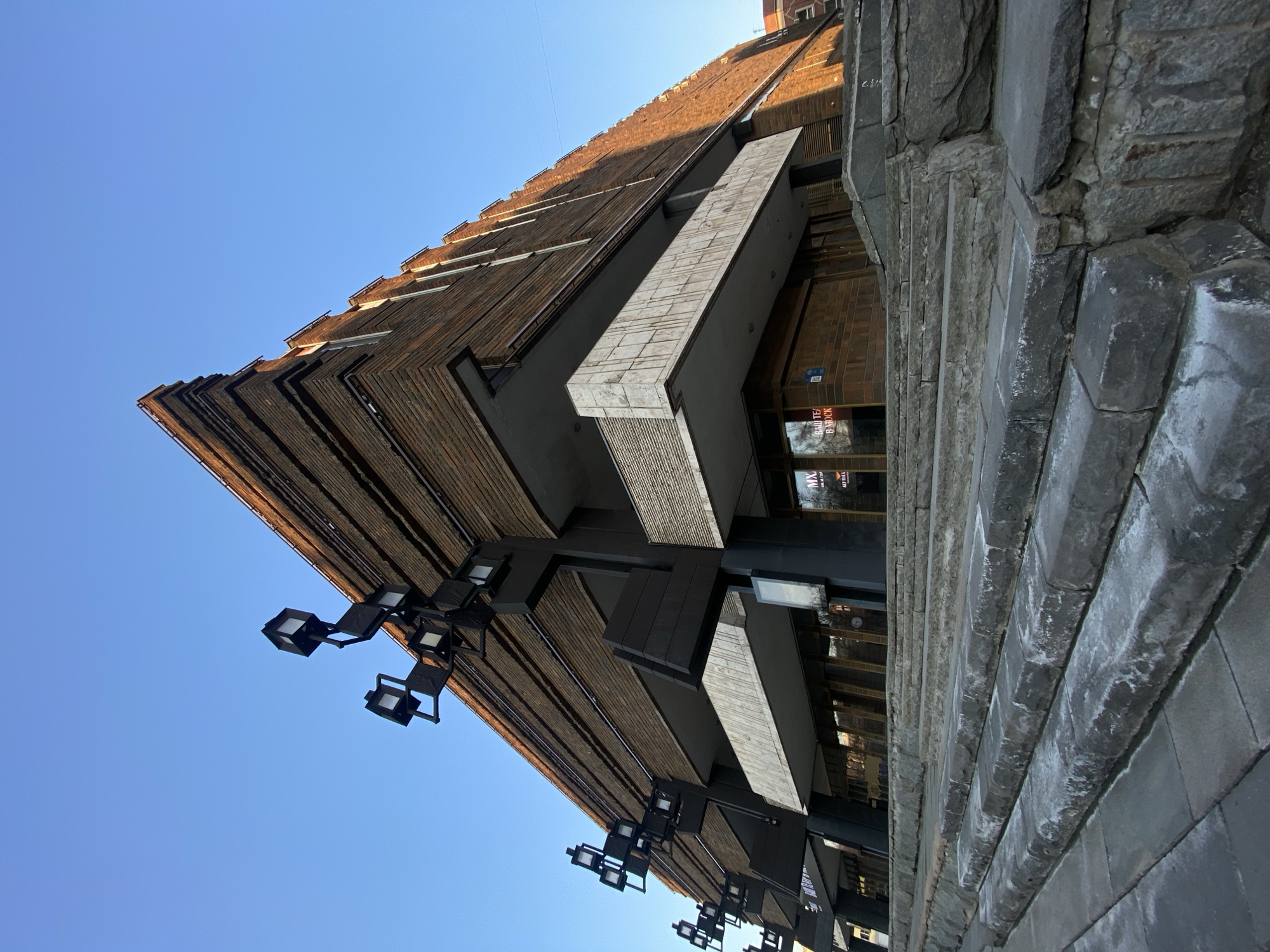 A low-angle exterior photograph depicts the Moscow Art Theatre Gorky (МХАТ им. М. Горького) located at Tverskoy Boulevard 22, Moscow, Russia. The multi-story building exhibits a Brutalist architectural style, characterized by a stepped, angular profile and a brown, textured facade composed of prominent horizontal bands, possibly brick or tile. Large, black, multi-armed light fixtures, each bearing multiple square luminaires, project from the upper sections of the facade on the left side. Recessed levels feature light-colored, textured concrete or stone balustrades. The ground level contains expansive reflective glass windows, with Russian text, including "МХАТ" and "ВАШ ТЕАТР В МОСКВЕ" (Your Theater in Moscow), visible in the reflections. A wide, multi-tiered staircase of rough, grey stone with visible weathering and white residue on the treads occupies the foreground, leading towards the building's entrance. The sky is clear and bright blue. No individuals are present in the scene.
