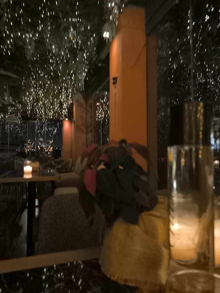 The image depicts a dim, ambient scene at an evening establishment in Palaio Faliro, Greece, likely a restaurant or lounge. The time of day is night, with the primary illumination coming from countless small, warm-toned string lights suspended above, possibly on trees or foliage, creating a sparkling, festive canopy.
In the immediate foreground, a dark, reflective tabletop, possibly marble, is visible. On the right, a tall, clear glass or bottle stands with reflections of the ambient lights. To its left, a textured yellow/brown object, possibly a woven bag or cushion, is partially obscured. Resting on what appears to be a chair or another surface are folded personal belongings, including dark clothes with a prominent red or pink section, and a brown item.
The midground features a prominent, tall, ochre or orange-colored pillar or wall section. Further into the scene, another table is visible with a lit candle, contributing to the intimate atmosphere. Glasses and perhaps other dinnerware are also on this table. Seating areas with upholstered chairs are faintly discernible around the tables.
The background reveals more of the sparkling lights, suggesting a large outdoor or semi-enclosed area. Reflections in glass panels imply a partition or wall, through which distant, blurred city lights or other illuminated structures can be seen. No people are clearly visible, but the setup indicates a space prepared for patrons.
The overall activity suggested is a relaxed evening out, such as dining or enjoying drinks. The ambiance is cozy and magical, attributed to the abundance of decorative lights. There is no visible text in the image.