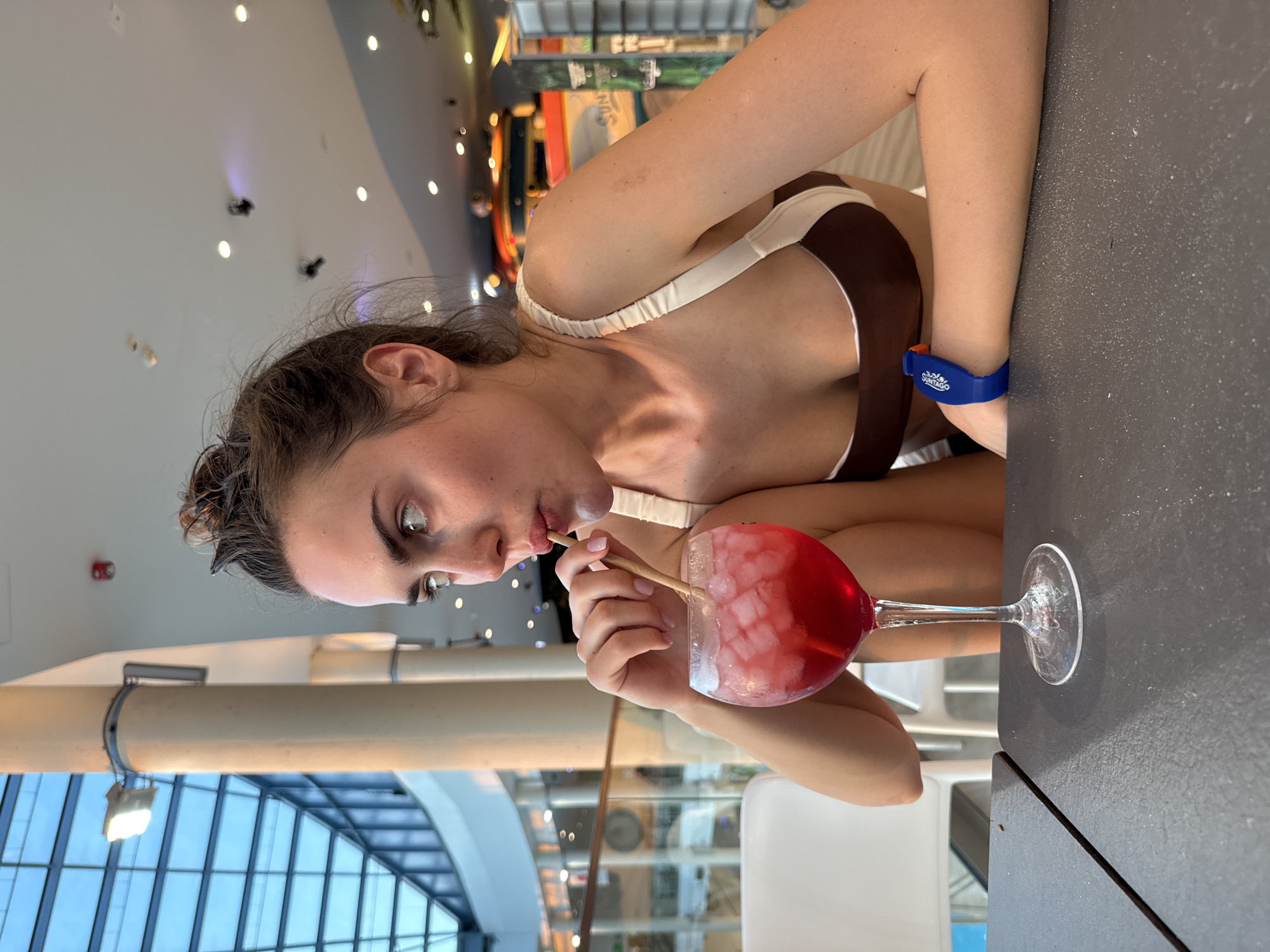 A woman with brown hair and a brown and white bikini top is drinking a red drink with ice from a tall glass using a straw at a gray table. She has a blue wristband on her left wrist. The location appears to be indoors near a large window with a view of a building with blue window panes. The city and specific location are unknown.