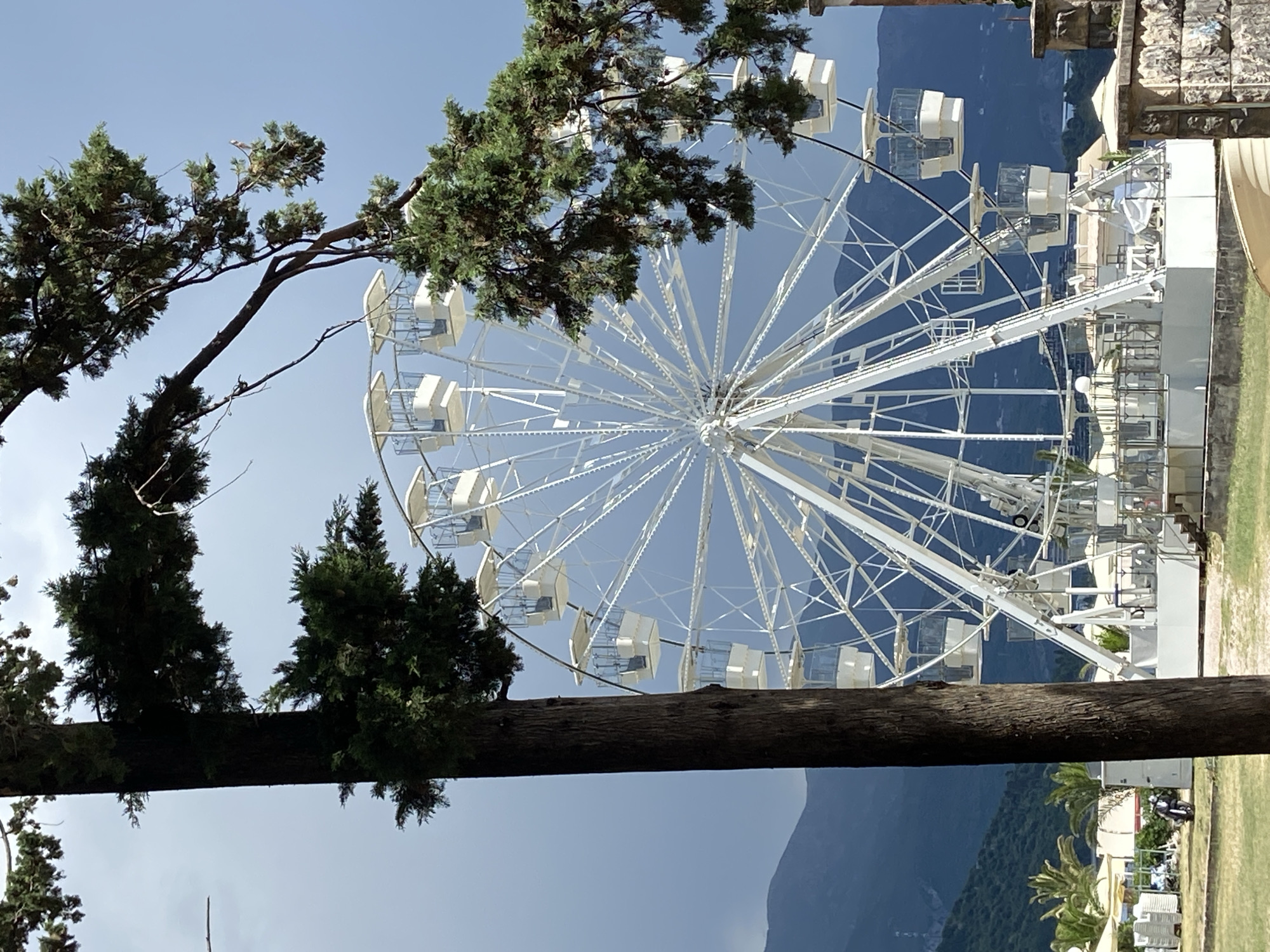 This image captures a white Ferris wheel against a clear blue sky, with mountains in the background. The Ferris wheel is a prominent object, featuring multiple enclosed white cabins. In the foreground, a large, dark tree trunk runs horizontally across the frame, with green foliage from two trees partially obscuring the Ferris wheel. The scene appears to be an outdoor amusement park or recreational area.
In the lower right portion of the image, there is a glimpse of a paved walkway with some parked chairs and a motorcycle. Further in the background, more structures and greenery are visible, suggesting a populated area or resort. The lighting indicates it is daytime, likely sunny due to the bright sky and clear visibility. The overall mood is relaxed and recreational.
The location is contextually identified as Budva, Montenegro, which aligns with the presence of a Ferris wheel and the mountainous, coastal landscape often found in the region. No text is visible in the image.