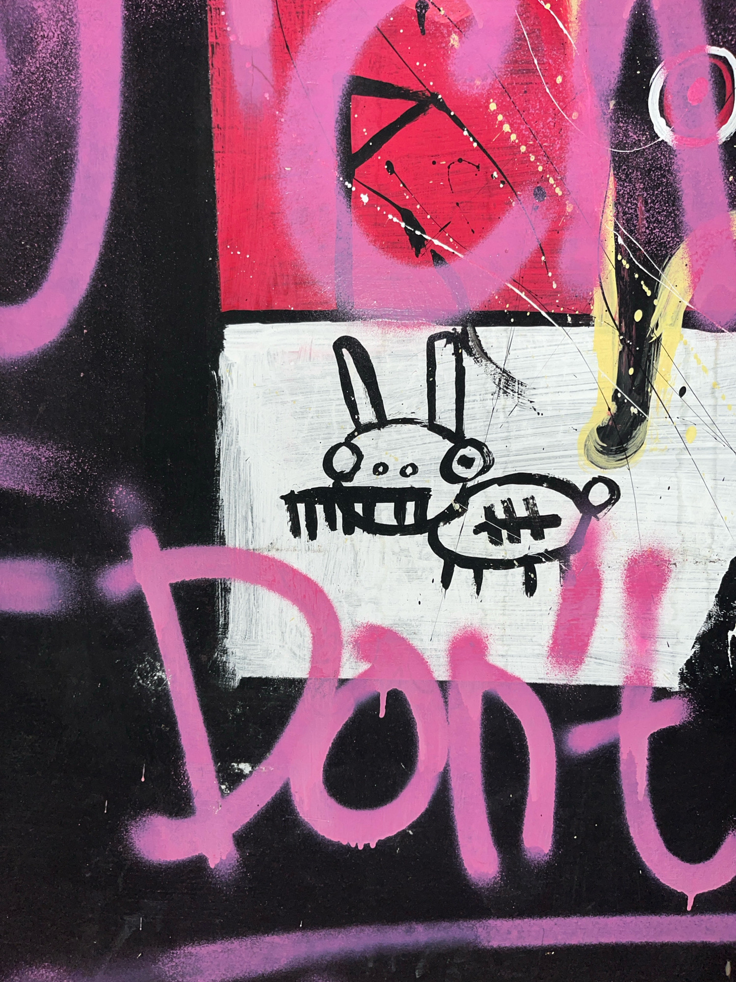 A close-up, full-frame, eye-level, outdoor, medium shot captures a piece of graffiti art on a vertical surface in Surakarta, Indonesia. The artwork features a mix of spray paint and hand-drawn elements. Dominating the foreground is a large, bold pink spray-painted word, partially obscured by the edge of the frame, that appears to spell "Don't". To the left of this word, large, indistinct pink graffiti shapes are visible against a black background. In the upper portion of the image, a section of red with black lines and pink spray paint is visible. Below this red section, on a white background, are two crudely drawn black figures, resembling a rabbit with ears and a simplified, somewhat pig-like creature with hash marks. The artwork exhibits signs of age and weathering, with drips and splatters of paint adding texture to the composition. The overall impression is one of urban street art.