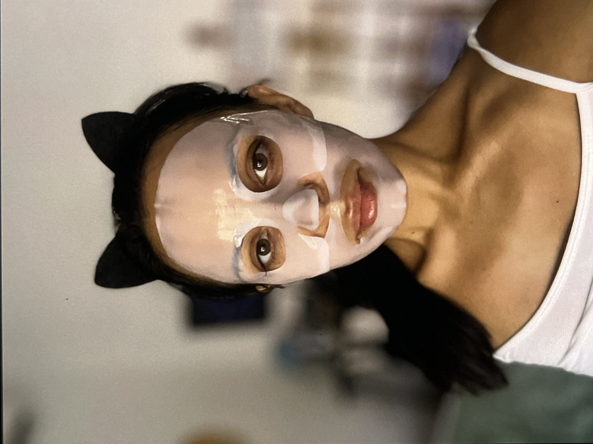 A woman wearing a white tank top and a black headband with cat ears is shown from the chest up, turned sideways. Her face is covered with a sheet mask. The background is blurred. The location in the city of Unknown, Indonesia is not discernible from the image.