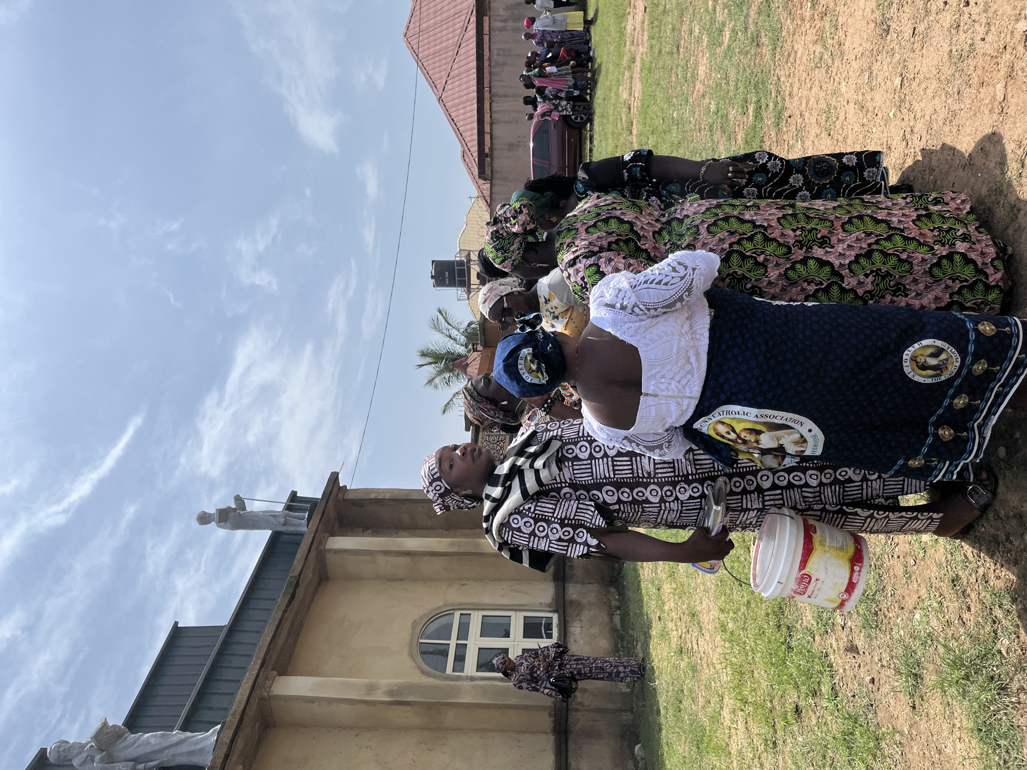 In Barakin Akawo, Nigeria, a group of women in traditional attire are gathered outdoors near a building with statues on its roof. The building appears to be a church, made of light-colored stucco with a prominent arched window. One woman in the foreground holds a red and white bucket. In the background, other women and children are visible, and a car is parked. The scene is set under a bright, partially cloudy sky.