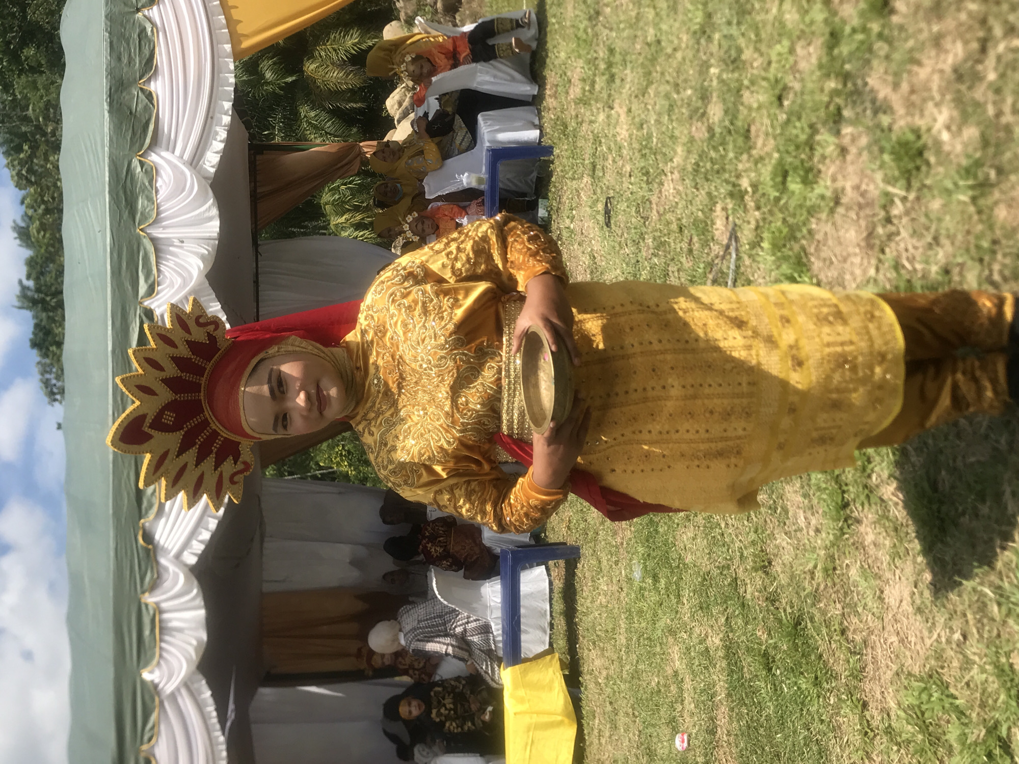 In Langkuta, Indonesia, a young woman stands in traditional yellow attire, adorned with intricate gold embroidery, holding a small brass bowl. She wears a golden headdress with red accents and a red veil draped over her shoulders. The backdrop features a green tent with white drapes, suggesting an outdoor event or celebration. Several individuals are seated in chairs behind her, some appearing to be children, also dressed in festive clothing. The ground is covered in grass, indicating an outdoor setting, possibly a park or a garden. The sky is visible with white clouds.