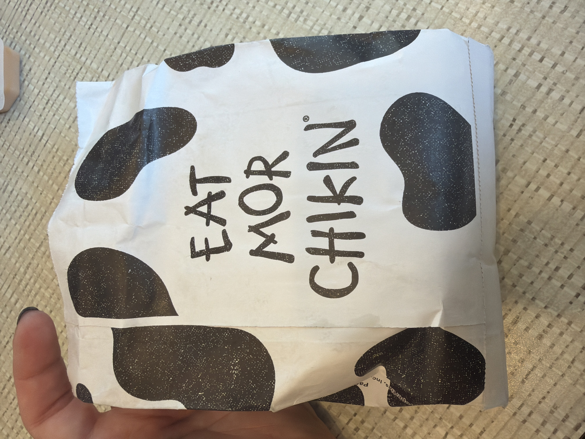 A hand is holding a white paper bag with a cow print pattern. The bag has black, irregular splotches with white speckles. The words "EAT MOR CHIKIN" are printed in bold, black, sans-serif font in all capital letters on the front of the bag. A small trademark symbol is visible to the right of the word "CHIKIN." The bag is wrinkled and appears to be crumpled at the bottom. The background is a woven textured surface, possibly a placemat or table. The image appears to be taken indoors, likely in a restaurant or at home.