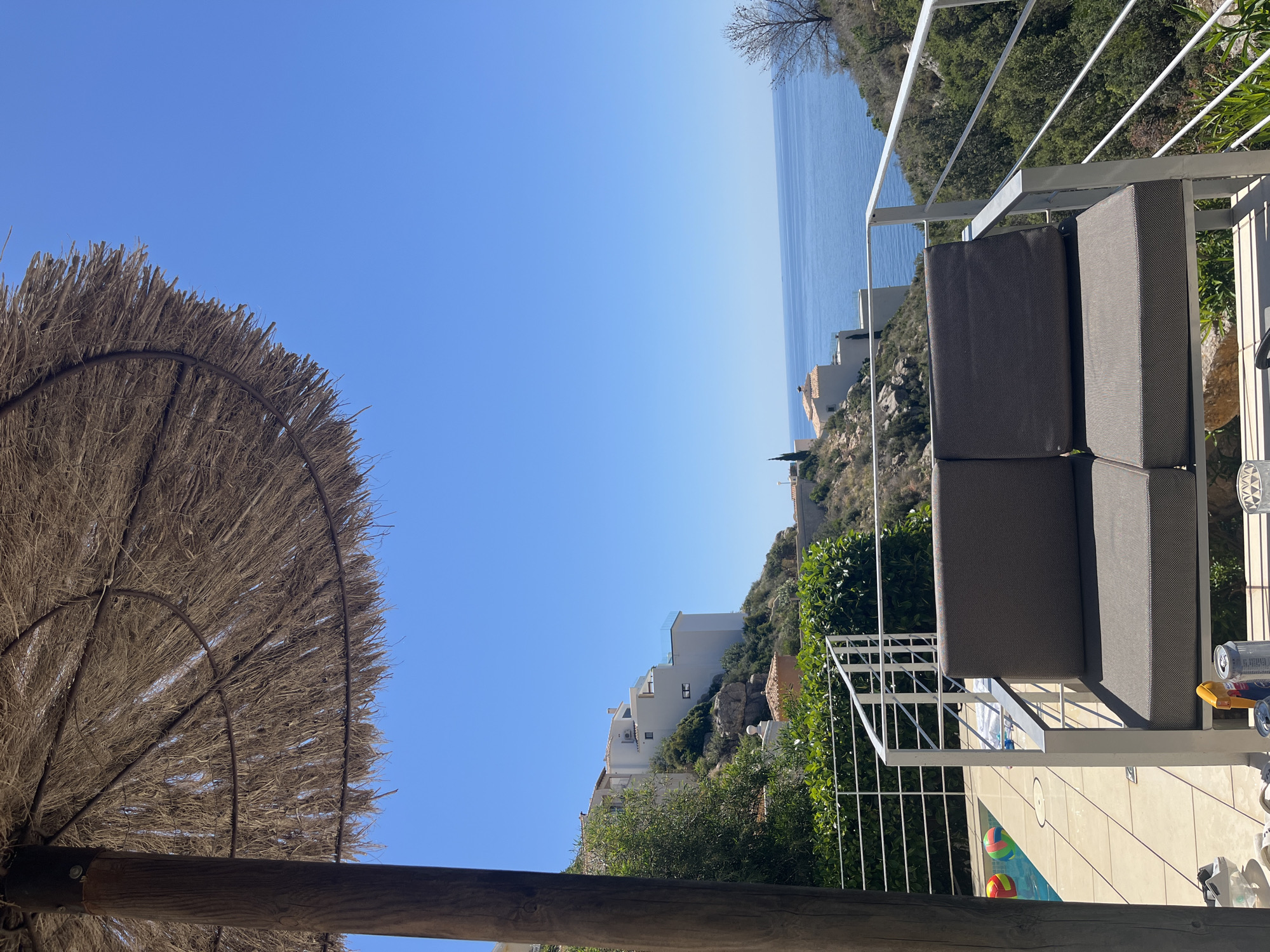 View from a terrace overlooking the Mediterranean Sea in El Poble Nou de Benitatxell, Spain. A thatched parasol is on the left. A gray outdoor sofa sits behind a white metal railing. Below is a swimming pool with inflatable balls. White buildings are on a hill, against a clear blue sky.