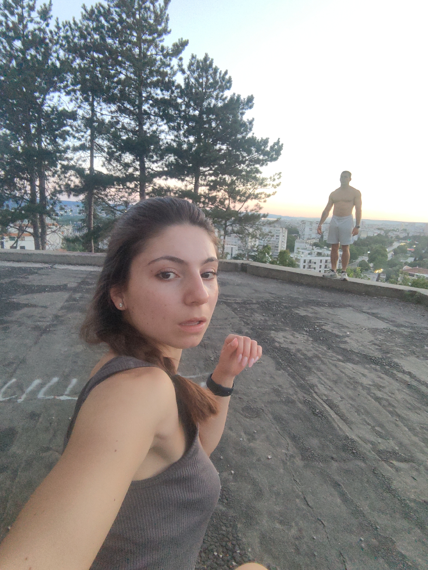 A selfie is taken by a young woman from a close-up, low-angle perspective, capturing her face and upper body. Her hair is dark brown and pulled back, with strands framing her face. She is wearing a grey tank top and a black fitness tracker on her left wrist. Her right hand is visible in the foreground, raised slightly, with neatly manicured fingernails. She appears to be looking at the camera with a neutral expression.
In the background, a shirtless man stands on a concrete ledge overlooking the city of Varna, Bulgaria. He is wearing light-colored shorts and athletic shoes. He is facing away from the camera, with his body in a relaxed stance. Behind him, the cityscape unfolds with numerous buildings and greenery, bathed in the soft light of what appears to be sunset or sunrise. Tall evergreen trees are visible on the left side of the frame, adding a natural element to the urban landscape. The ground in the foreground is a rough, grey concrete surface with some white markings. The overall scene suggests an outdoor setting, possibly an elevated viewpoint or park within the city.