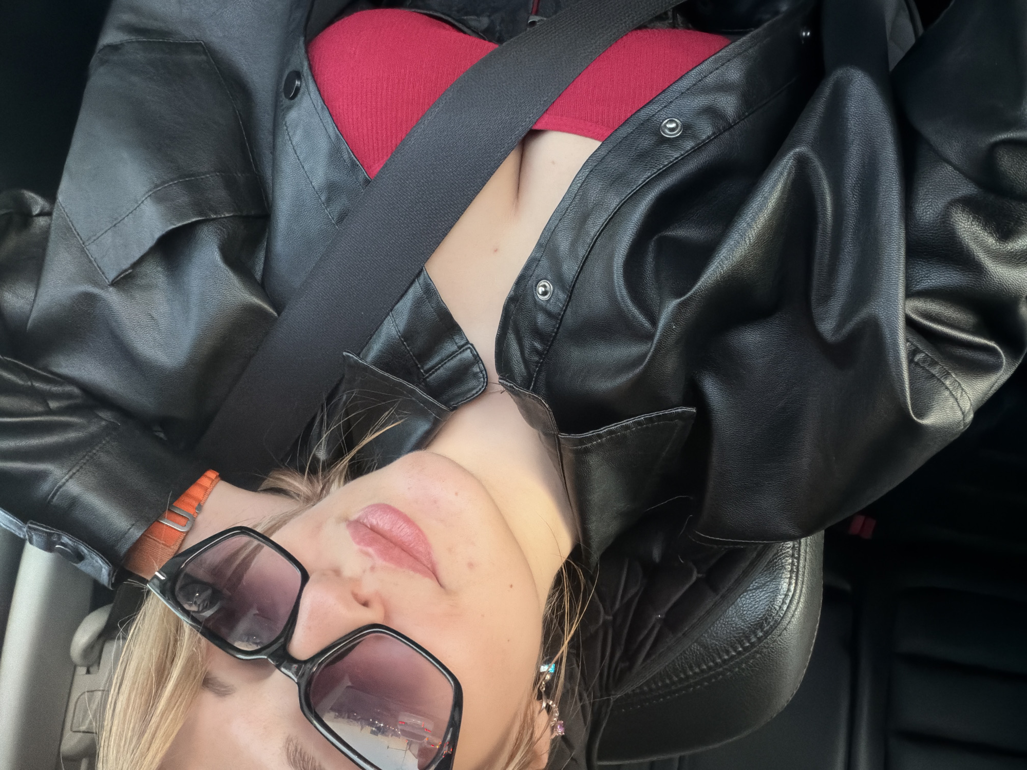 A woman is shown in an indoor setting, likely inside a vehicle. She is wearing a red top, a black leather jacket, and large black-framed sunglasses. A seatbelt is visible across her chest. The image seems to be a selfie or taken at a low angle, showing her face and upper body. The location of the photo is unknown and cannot be ascertained based on the image alone.