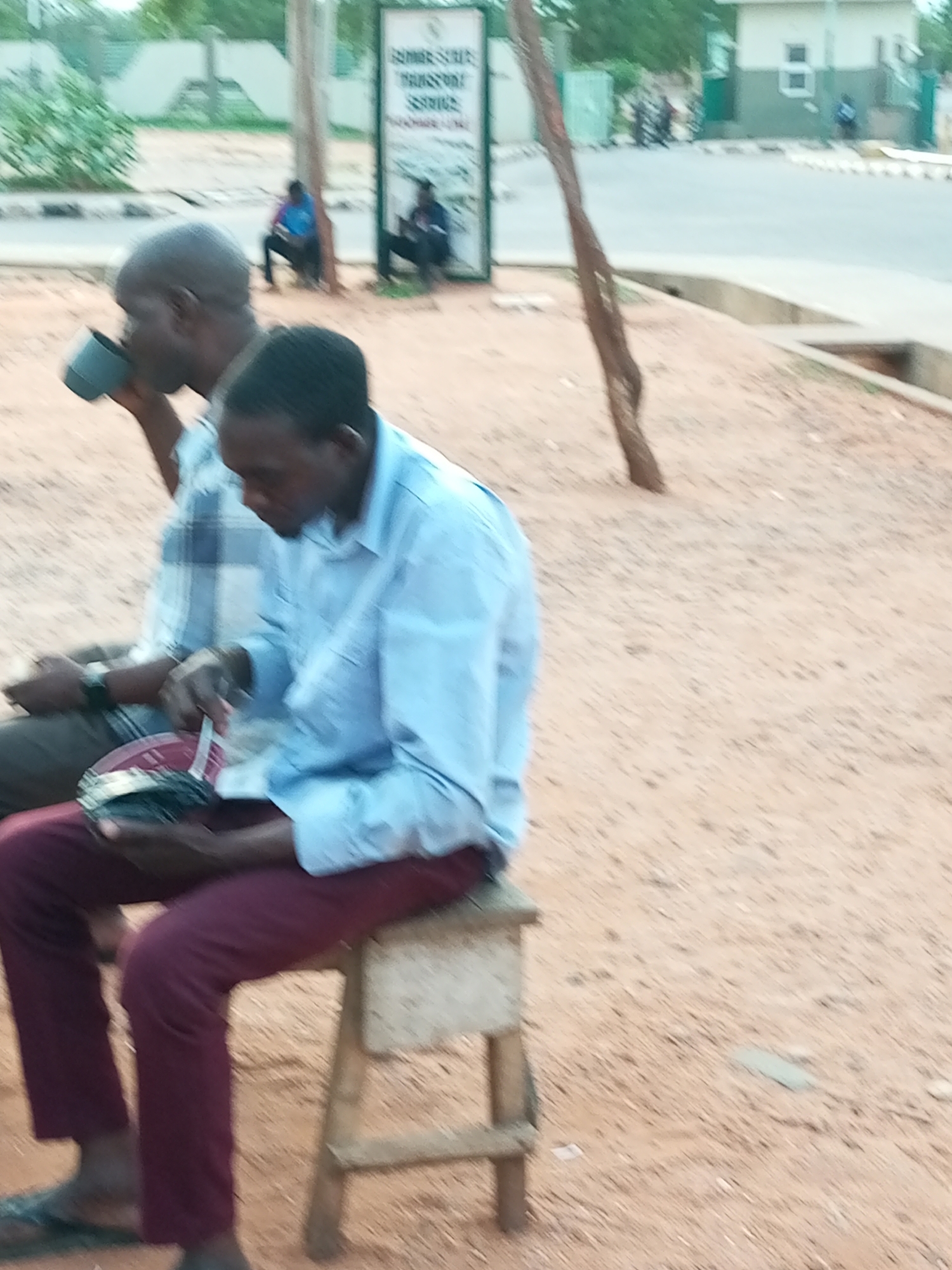 In Gombe, Nigeria, two men are seated outdoors on a sandy ground. The man in the foreground, wearing a blue collared shirt and maroon pants, is looking down at a red and black object he is holding in his hands. He appears to be stirring or examining its contents. To his left, another man, wearing a checkered shirt and dark pants, is holding a gray cup to his mouth, seemingly drinking. Behind them, a sign with text is visible on a pole, and in the background, there are buildings and a road with a few other people in the distance. The overall scene suggests a casual outdoor gathering or waiting area.