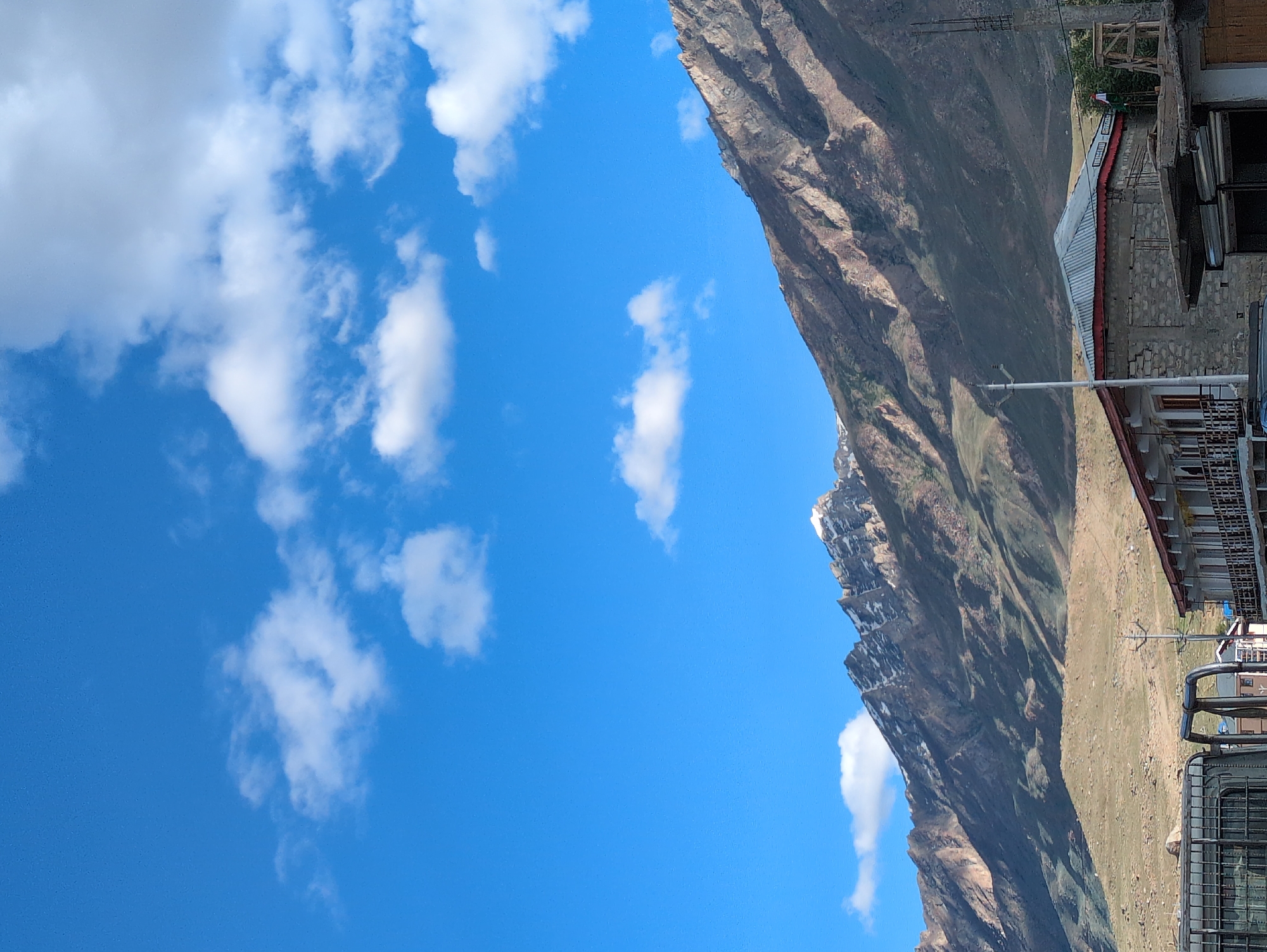 The image depicts a mountainous landscape under a bright blue sky dotted with white clouds. In the foreground, there is a building with a red-trimmed roof and stone walls. A metal fence and parts of a vehicle are visible on the left. A utility pole stands near the building. The middle ground shows a barren, sloping hillside leading up to a majestic mountain range, whose peaks are dusted with snow. The mountain slopes are characterized by dry vegetation and shadows. The scene is likely a street view in or near the city of Padum, India, given the architecture and mountainous terrain.