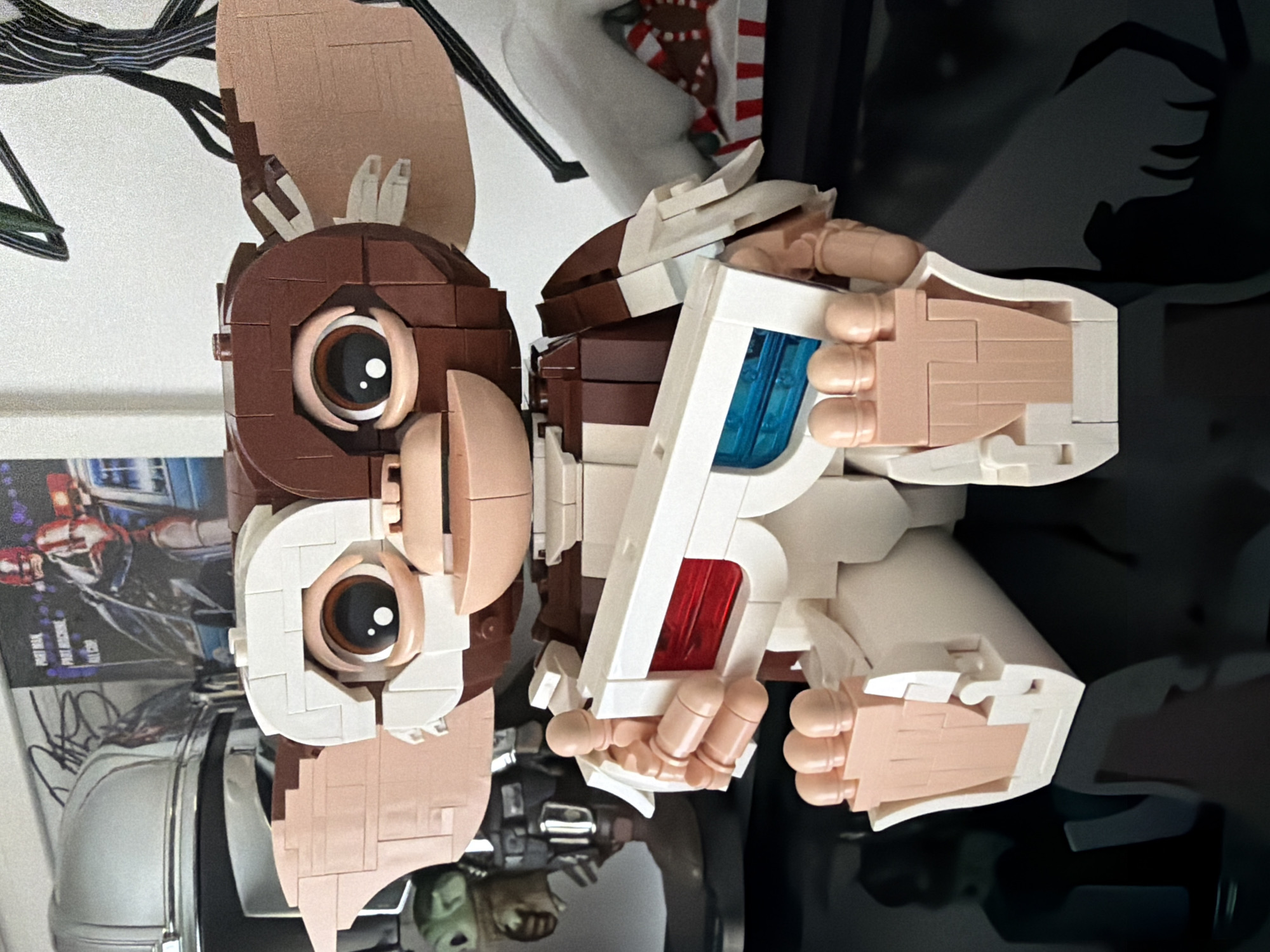 A Lego model of Gizmo from the movie "Gremlins" is displayed on a reflective surface, likely a shelf or table. Gizmo is holding a pair of 3D glasses with blue and red lenses. In the background are various objects: a framed poster, a white sculpture, and some other indistinct items. The location of Pétange, Luxembourg, cannot be determined from the visual information in this image.