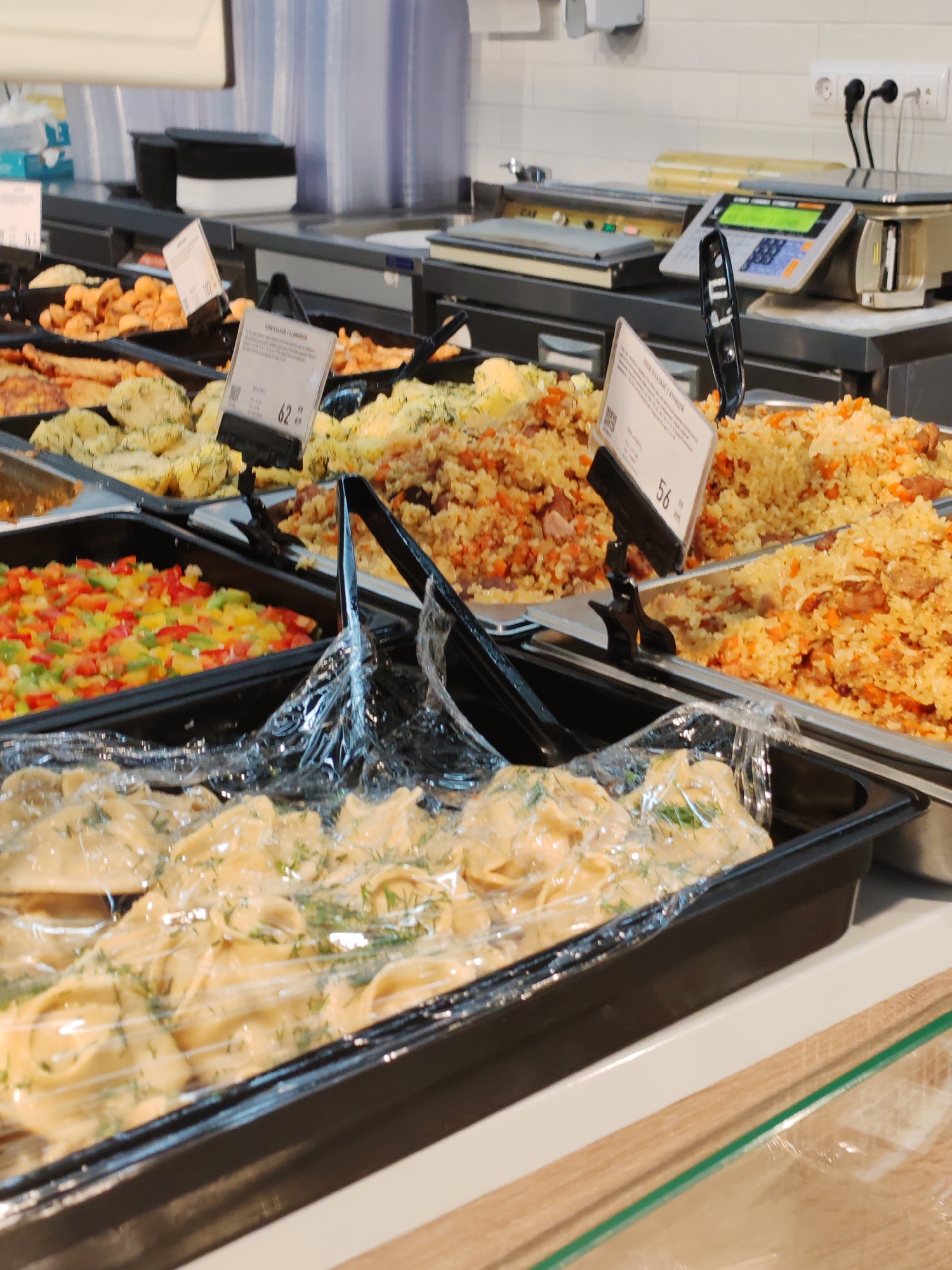 The image shows a food display case in a supermarket in Komsomolsk-on-Amur, Russia. The display case contains various prepared foods, including rice pilaf, chopped vegetables, and dumplings. Price tags are visible in front of some of the dishes. In the background, a commercial scale and a checkout counter are visible. The lighting is bright and artificial, typical of a retail environment.