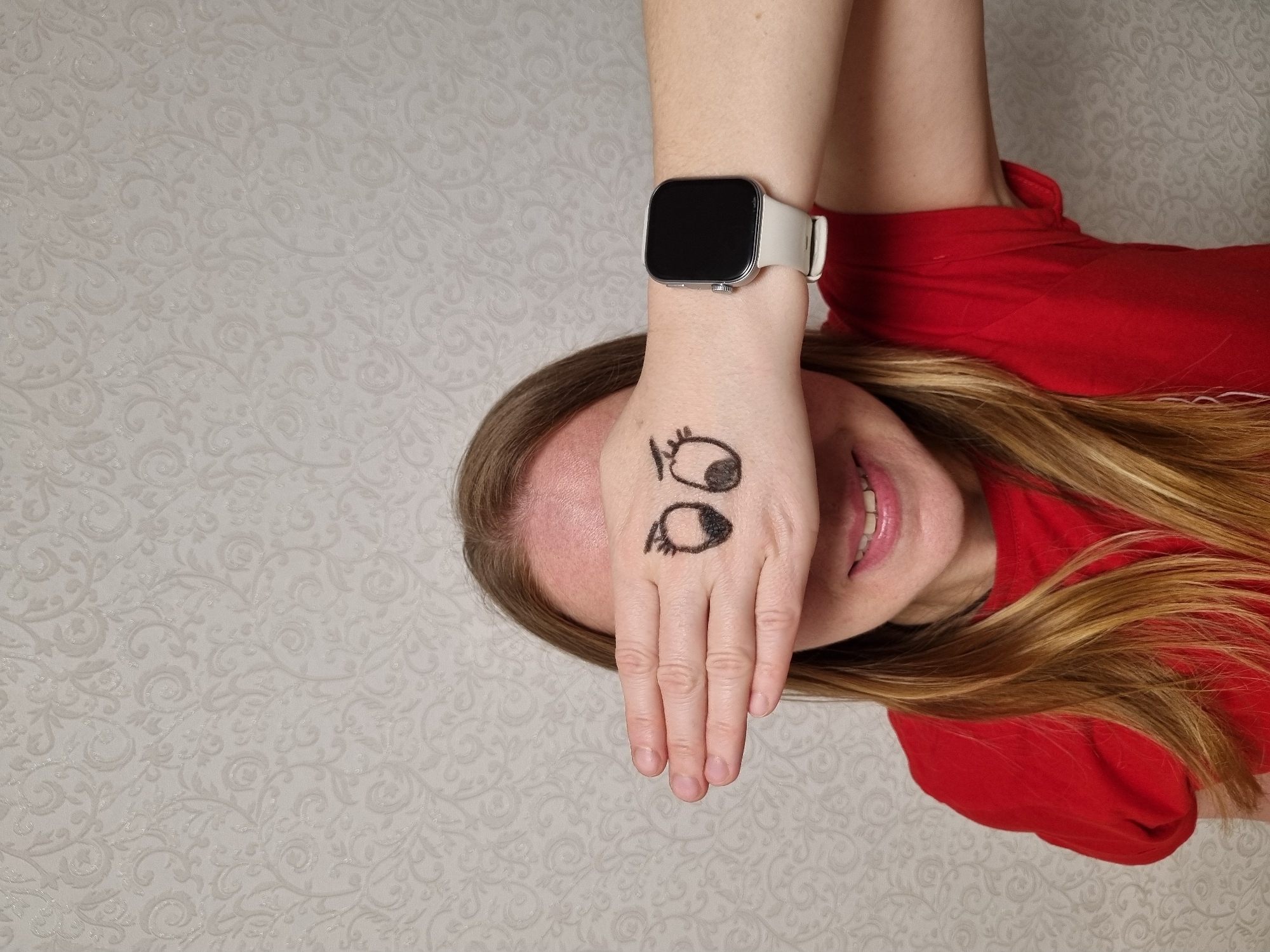 A woman is seen from a low-angle perspective, seemingly lying on her side with her body oriented towards the right of the frame. She is wearing a red shirt and has light brown or blonde hair. Her right hand is held up in front of her face, obscuring her eyes. On the back of her hand, there are cartoonish drawings of eyes. She also has a smartwatch on her right wrist. The background is a patterned off-white or light beige wall. The location is presumably Chaikovsky, Russia.