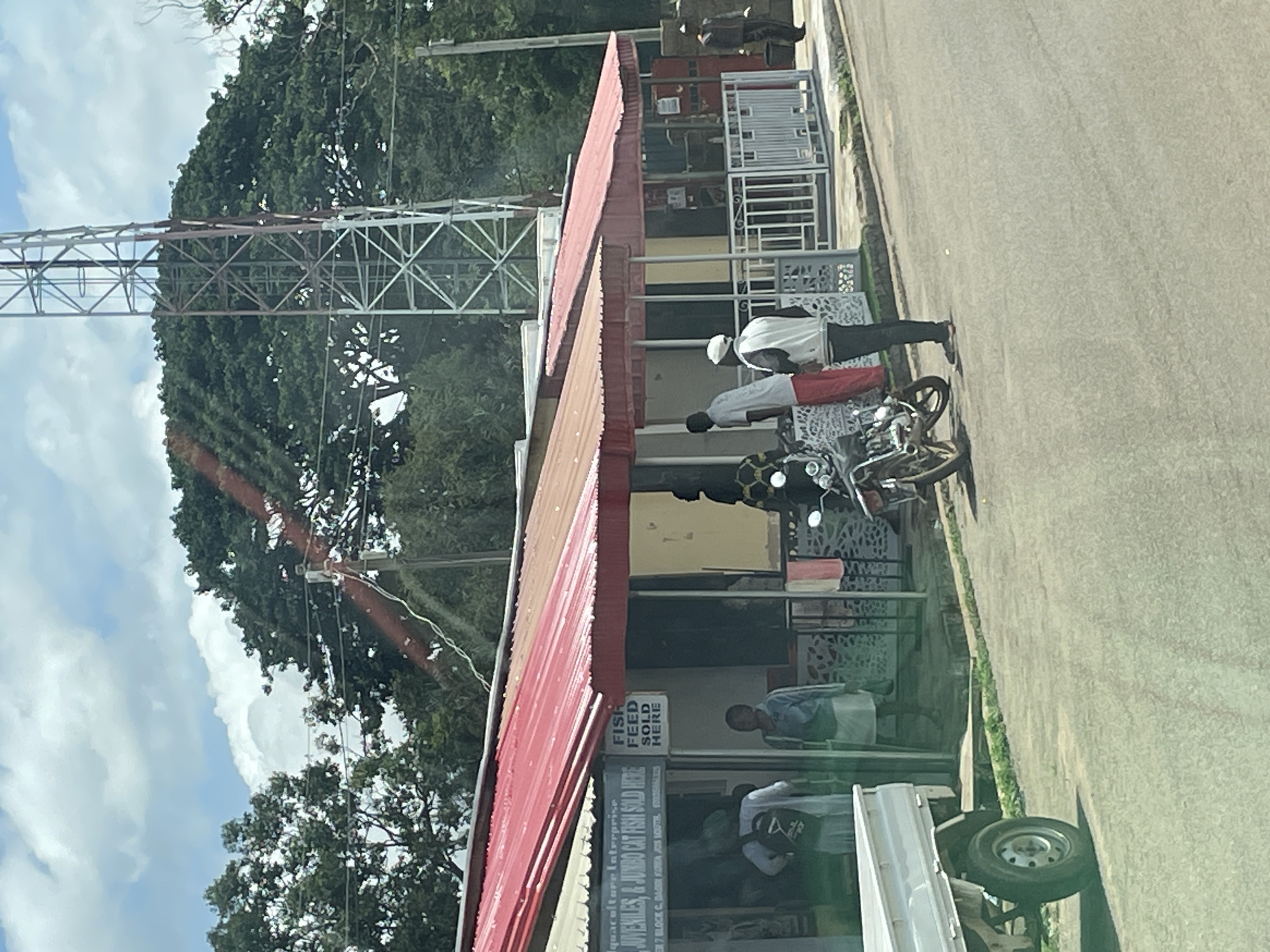 The image depicts a street scene in Jos, Nigeria, with a modest commercial building featuring a red corrugated metal roof and awnings. Several individuals are visible outside the building. Two people stand near a white pickup truck parked on the left side of the frame. A motorcycle is parked in front of the building to the right, with three people standing near it. Two individuals are visible inside the building through an open doorway. A white sign on the building reads "FISH FEED SOLD HERE". The environment includes a paved road in the foreground, a grassy area between the road and the building, and lush green trees in the background. A large telecommunications tower is prominent in the upper right portion of the image. The sky is partially cloudy.