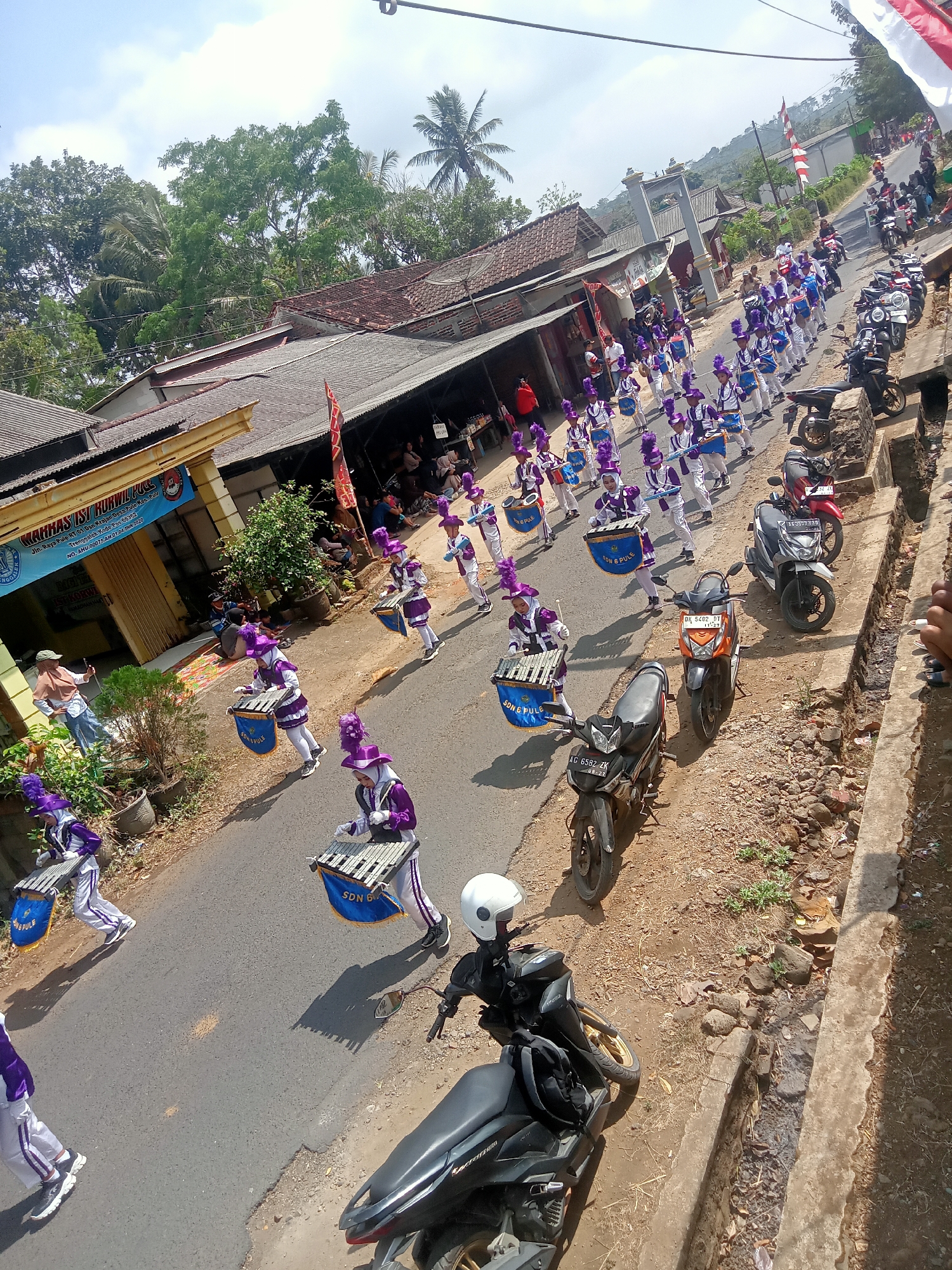In the city of Pule, Indonesia, on a sunlit day with a partly cloudy sky, a marching band parade is underway on a paved street lined with buildings and trees. The band members, predominantly young individuals in matching purple and white uniforms adorned with purple hats and plumes, are playing various instruments including drums and xylophones. The street is populated with spectators, some of whom are seated on the side, and several motorcycles are parked or moving along the roadside. The buildings, characterized by their tiled roofs and open facades, suggest a village or small town setting. The parade appears to be in motion, with the band members marching in formation, creating a lively procession. The ground is a mix of paved road and dusty dirt with some vegetation and rocks on the side.
