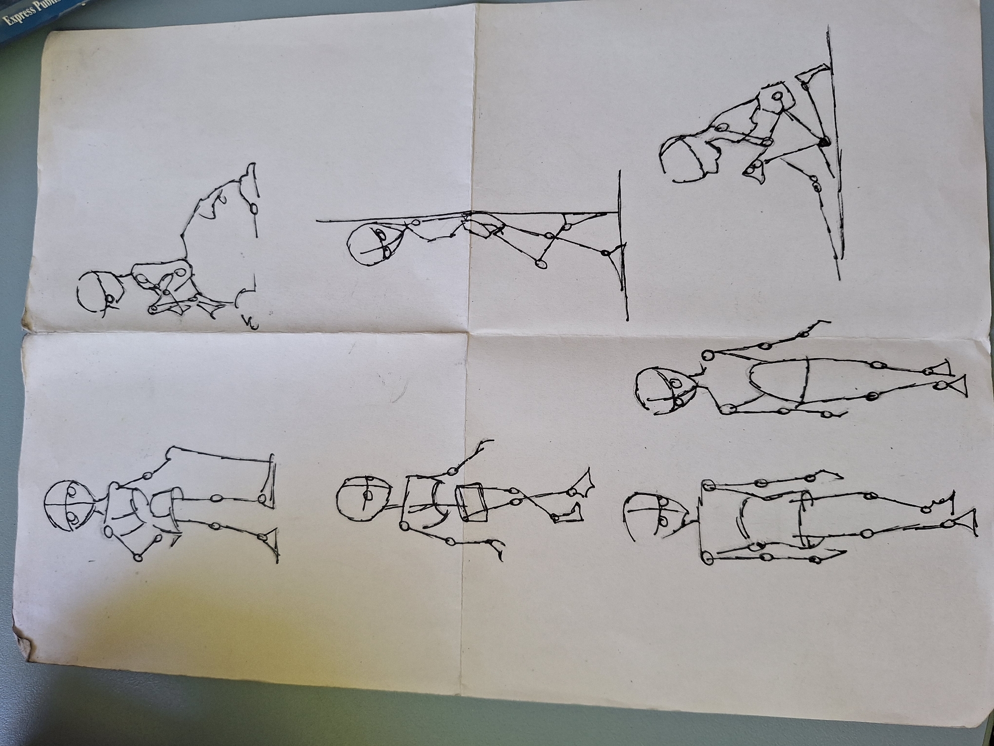 The image displays a sheet of paper with six pen-and-ink drawings of human figures. The drawings are simple, skeletal in nature, and appear to be studies of human anatomy and pose.
In the top left, a figure stands with arms akimbo, one leg slightly forward.
In the top right, a figure is seated on an amorphous lump, holding something in its hands.
In the middle left, a figure stands in a walking pose, with one leg extended forward.
In the middle right, a figure leans against a vertical line, appearing to be looking downwards.
In the bottom left, two figures stand side-by-side, facing forward. The left figure has its arms at its sides, and the right figure has its arms slightly out.
In the bottom right, a figure is kneeling on one knee, with its hands resting on its knees or the ground.
The paper itself is creased, suggesting it has been folded. The background is a plain, light-colored surface, possibly a table or wall. The lighting suggests it is an indoor environment. There are no explicit urban or natural elements present in the drawing itself, nor any specific geographical markers that would indicate a location in Varna, Bulgaria. The drawings are abstract studies and do not depict a recognizable scene from any particular city.