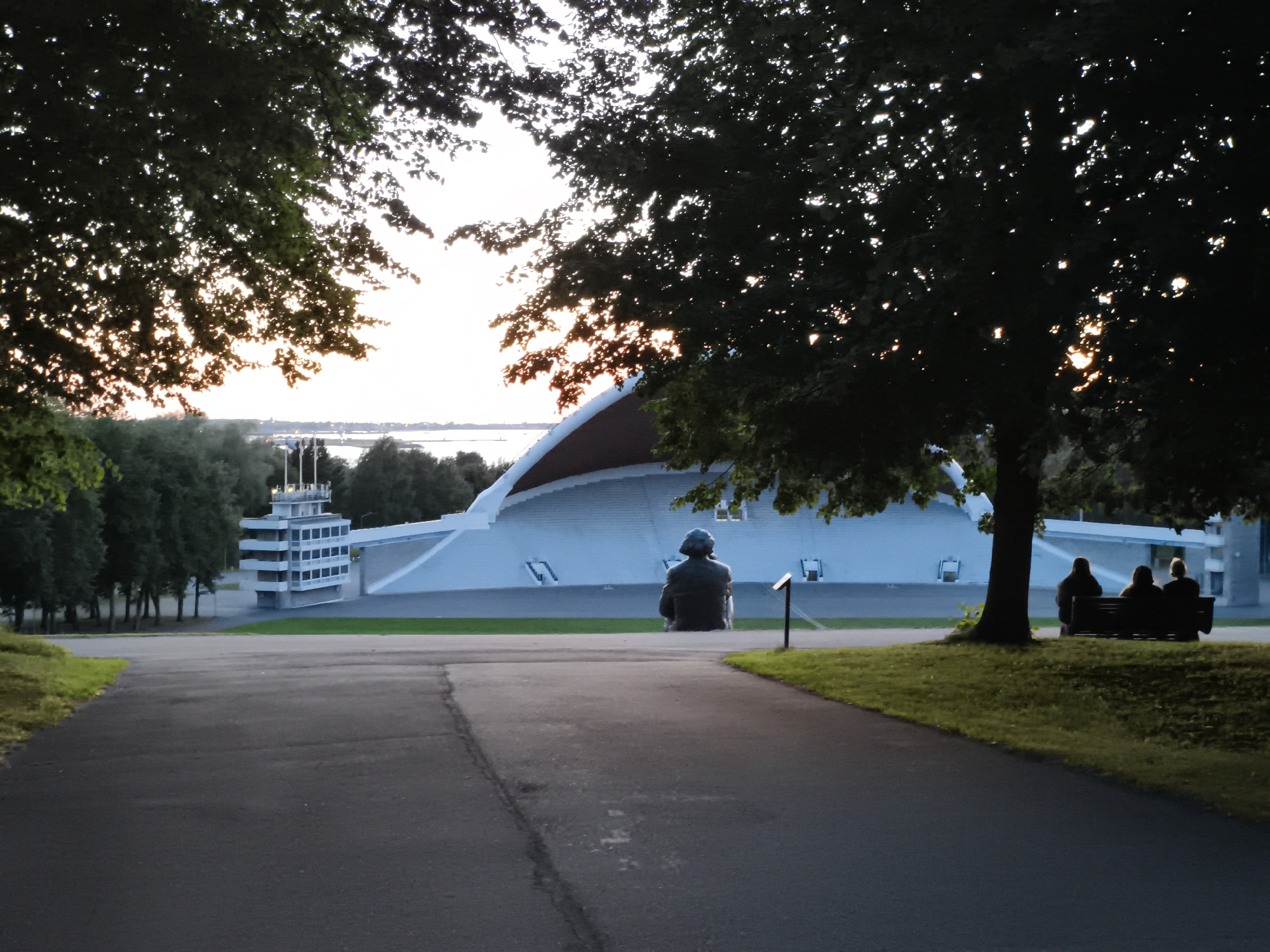 The image shows a view of the Tallinn Song Festival Grounds in Tallinn, Estonia. In the foreground, a paved path leads towards a large, white, amphitheater-like structure. To the left of the path, there is a grassy slope with trees. To the right of the path, there is a grassy area with several trees and a bench.
In the middle ground, a solitary statue of a seated figure is positioned on the grass, facing the song festival stage. Further to the right, on the bench, three figures are seated, silhouetted against the background. Behind them, the large white song festival stage dominates the scene, with a distinctive curved roof. To the left of the stage, a multi-story white building with many windows and a tower is visible.
The image is taken during what appears to be late afternoon or early evening, with the sun setting behind the trees, casting a warm glow through the leaves and creating dappled light. The sky is a bright, hazy white. The overall impression is one of a peaceful, public outdoor space.