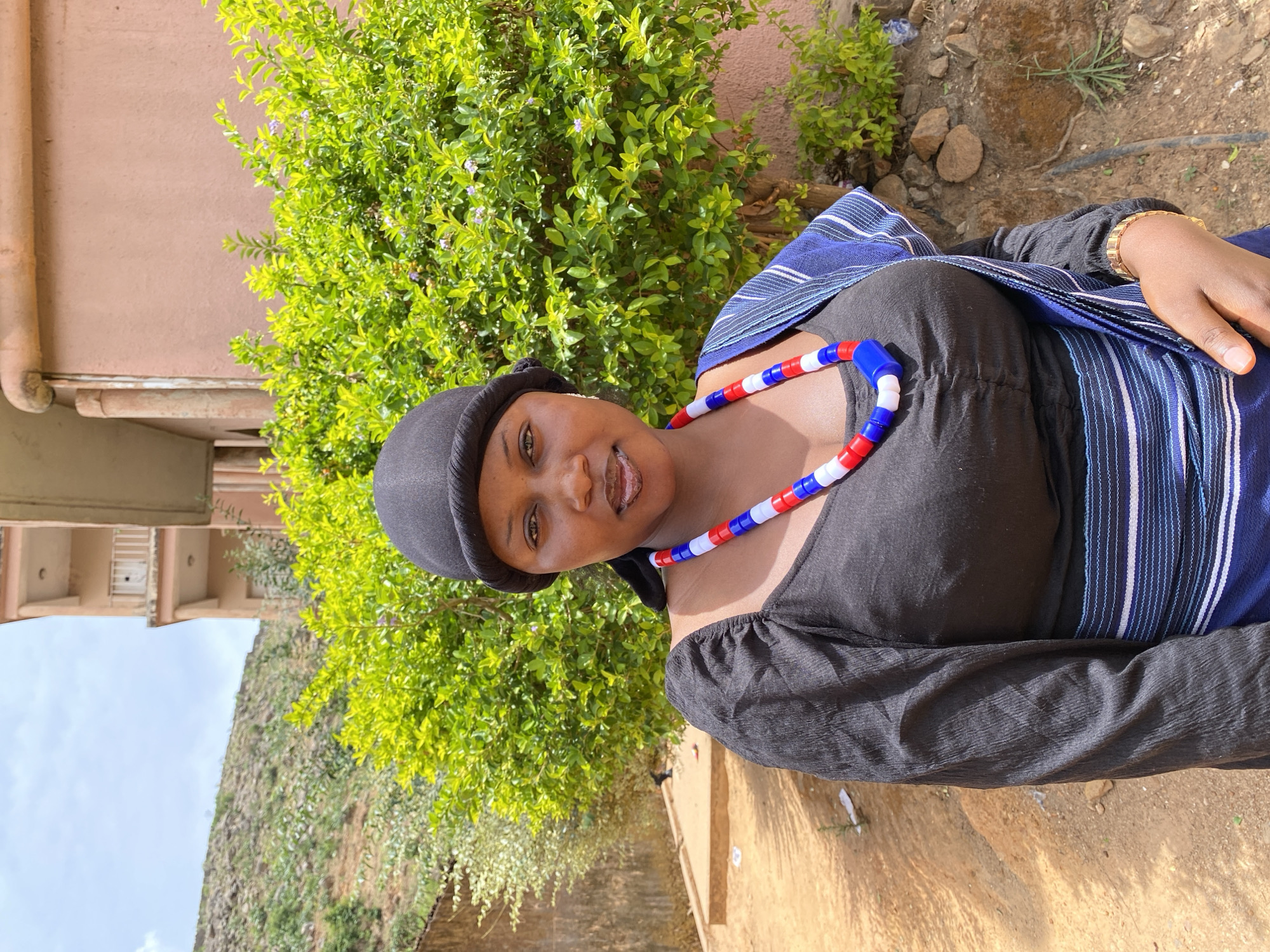 A woman wearing a black top, black hat, and a blue and white striped cloth over her shoulder stands outside. She has a necklace of red, white, and blue beads. Behind her is a green bush and a building with a pink exterior. The location is Gwong, Nigeria.