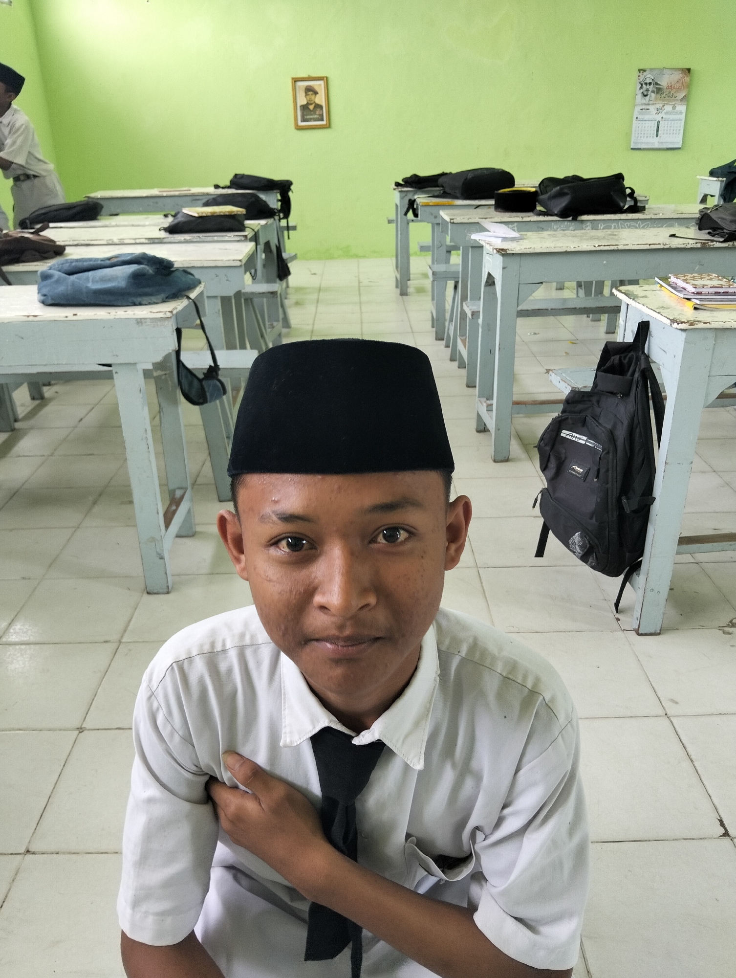 The image shows a young male, likely a student, wearing a white collared shirt, a black tie, and a black peci cap, positioned in the foreground. He is seated on the floor of a classroom in Kranji, Indonesia. The classroom features rows of light blue wooden desks with white tops, some of which have bags and books on them. The walls are painted a light green, and there are a framed portrait and a calendar hanging on the back wall. The floor is tiled in a light color. A black backpack is hanging on the leg of a desk behind the student. The student is looking directly at the camera with his right hand touching his chest. The overall scene depicts a typical classroom environment.
