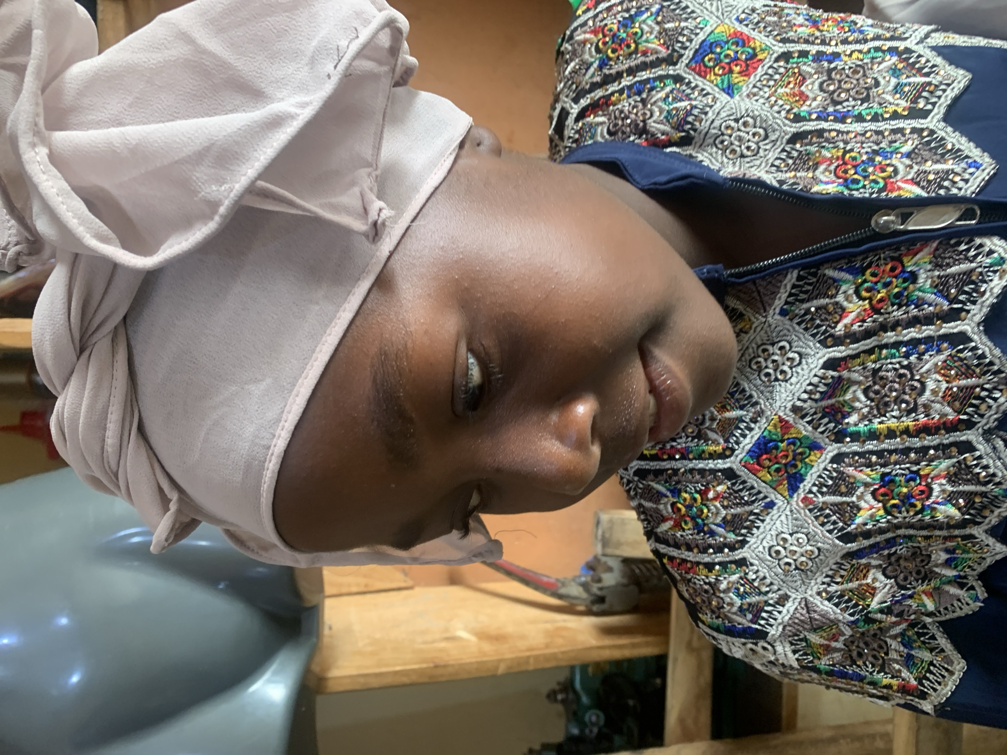 This media file is a close-up photograph of a young person wearing a light pink headscarf and a blue garment with intricate colorful embroidery. The person's head is tilted to the side, and their face is partially visible, showing their eye and lips. The background is blurred, but appears to be an indoor setting with wooden shelves and some equipment. The lighting suggests it could be daytime. The image is oriented sideways, with the person's head pointing towards the top of the frame. The embroidery on the garment features geometric patterns with small beads and colorful threads in shades of red, blue, green, yellow, and white. The overall impression is that of a portrait, focusing on the person's attire and facial features. The location context is Jos, Nigeria.