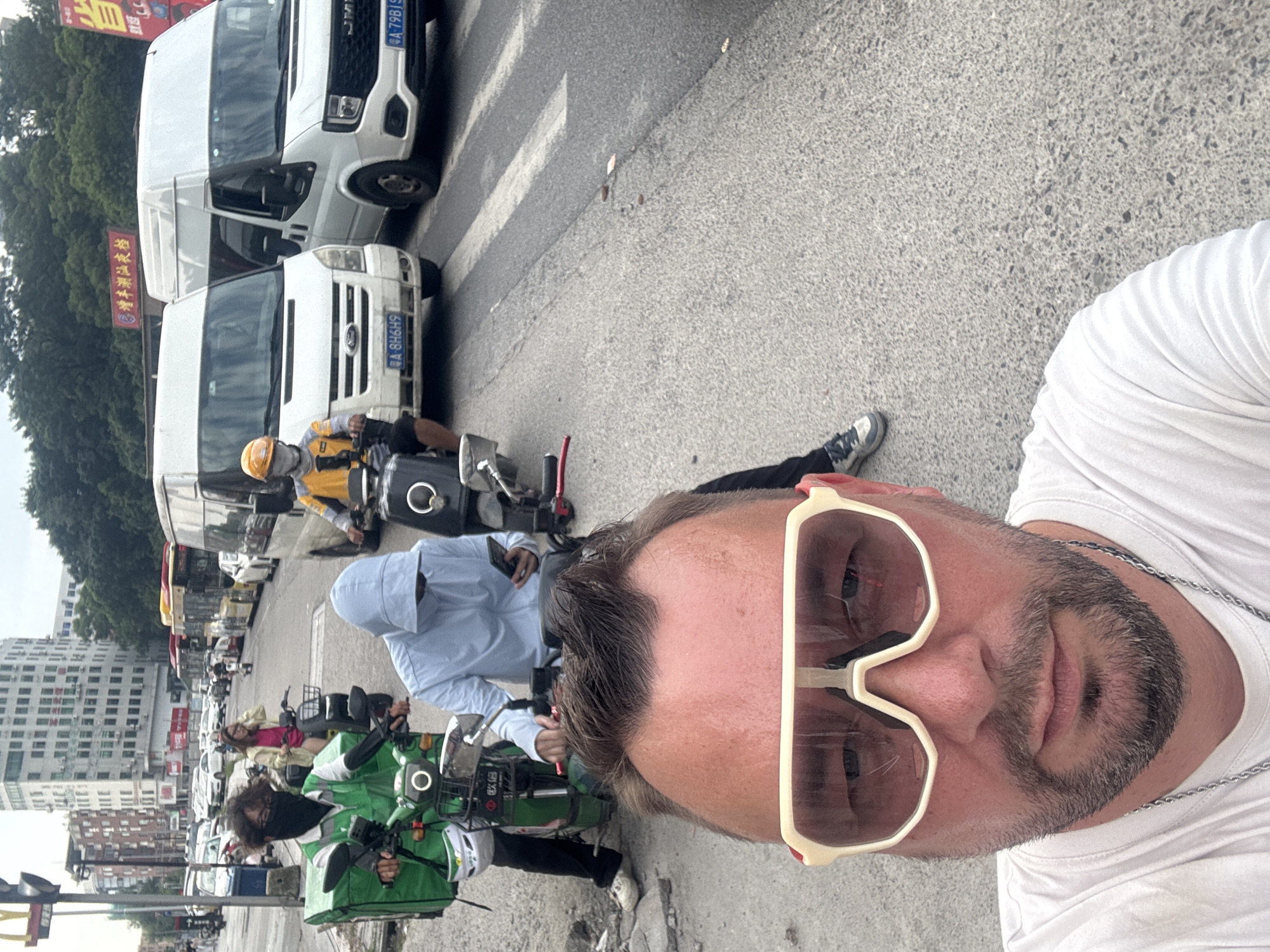 In the city of Nanhai District, China, a man wearing a white t-shirt and large white-framed sunglasses, with a beard and wearing a silver chain, takes a selfie on a street. Behind him, several people on motorcycles are visible. One individual, wearing a green vest and a black face mask, rides a green motorcycle with a large green delivery box. Another person in a light blue hooded jacket is looking at their phone while riding a motorcycle. Further back, a person in a yellow shirt and helmet is also on a motorcycle. White vans are parked on the side of the road, and a busy street with other vehicles and buildings can be seen in the background.