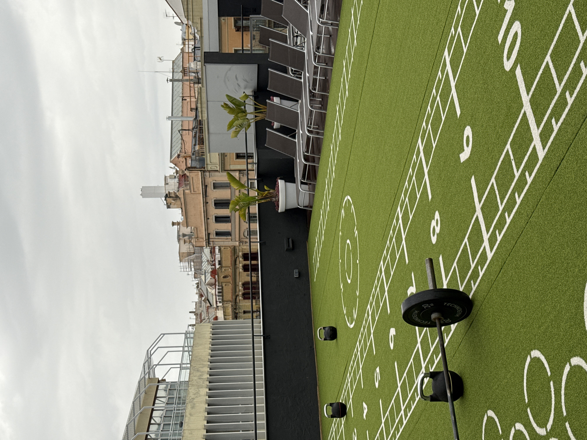 A rooftop outdoor gym in Barcelona, Spain is shown. The gym's floor is green artificial turf with white markings and numbers reminiscent of a football or exercise field. Exercise equipment, including a barbell with weight plates and kettlebells, is visible. In the background, the city's architecture is visible. There are buildings of different heights and styles with cloudy skies above them. Some lounge chairs are stacked along the side of the rooftop.