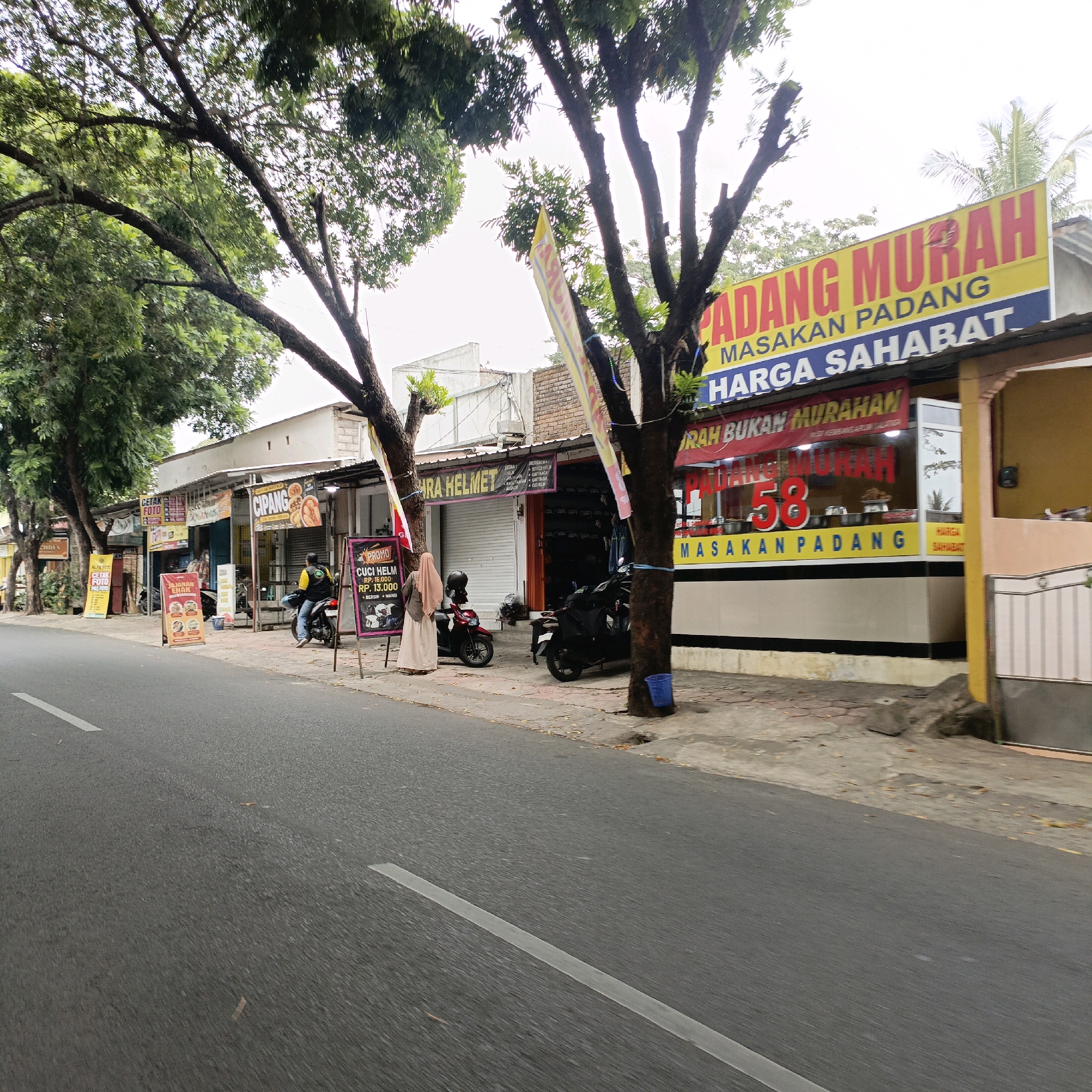 A street scene in Salatiga, Indonesia, shows a row of small businesses along the side of a paved road with white lane markings. The businesses include a "PADANG MURAH" restaurant with a bright yellow sign advertising "MASAKAN PADANG HARGA SAHABAT" (Cheap Padang Cuisine, Friendly Prices) and a red banner stating "MURAH BUKAN MURAHAN" (Cheap Not Shoddy). Another shop is marked "CIPANG" with an image of food. A shop selling "CUCI HELM" (Helmet Wash) has a promotional sign. In front of the shops, a man in a yellow shirt and black vest is seated on a motorcycle. A woman in a headscarf and long skirt stands near a motorcycle and a sign. Several trees with dense foliage line the street, casting shadows. The sky is overcast.