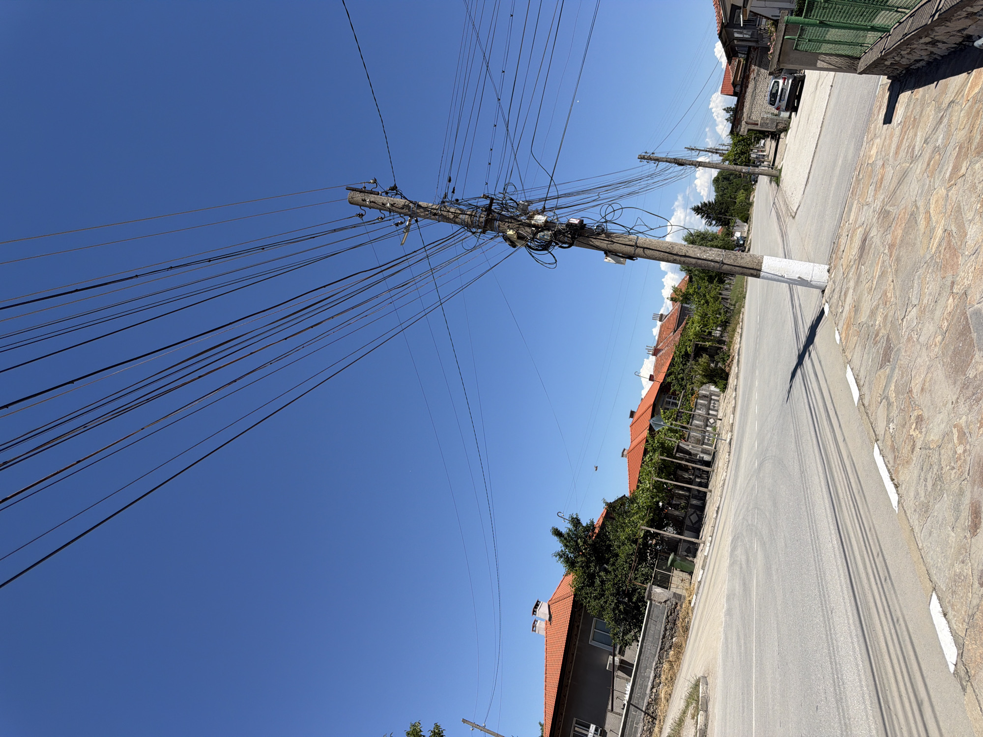 The image depicts a street scene in the city of Zlatitrap, Bulgaria. A prominent concrete utility pole, laden with a dense network of overhead electrical and telecommunication wires, stands near the foreground, leaning slightly to the right. The pole is painted white from its base to about a third of its height. To the left of the pole, a paved road curves, with tire marks visible on its surface. The road is bordered by a sidewalk made of irregular stone tiles.
In the background, several residential buildings with distinctive orange tiled roofs are visible, interspersed with trees and vegetation. A stone wall runs along the side of one property. A silver car is parked on the right side of the road, near a building. The sky is a clear, bright blue, indicating a sunny day. The scene appears to be a quiet, residential area.