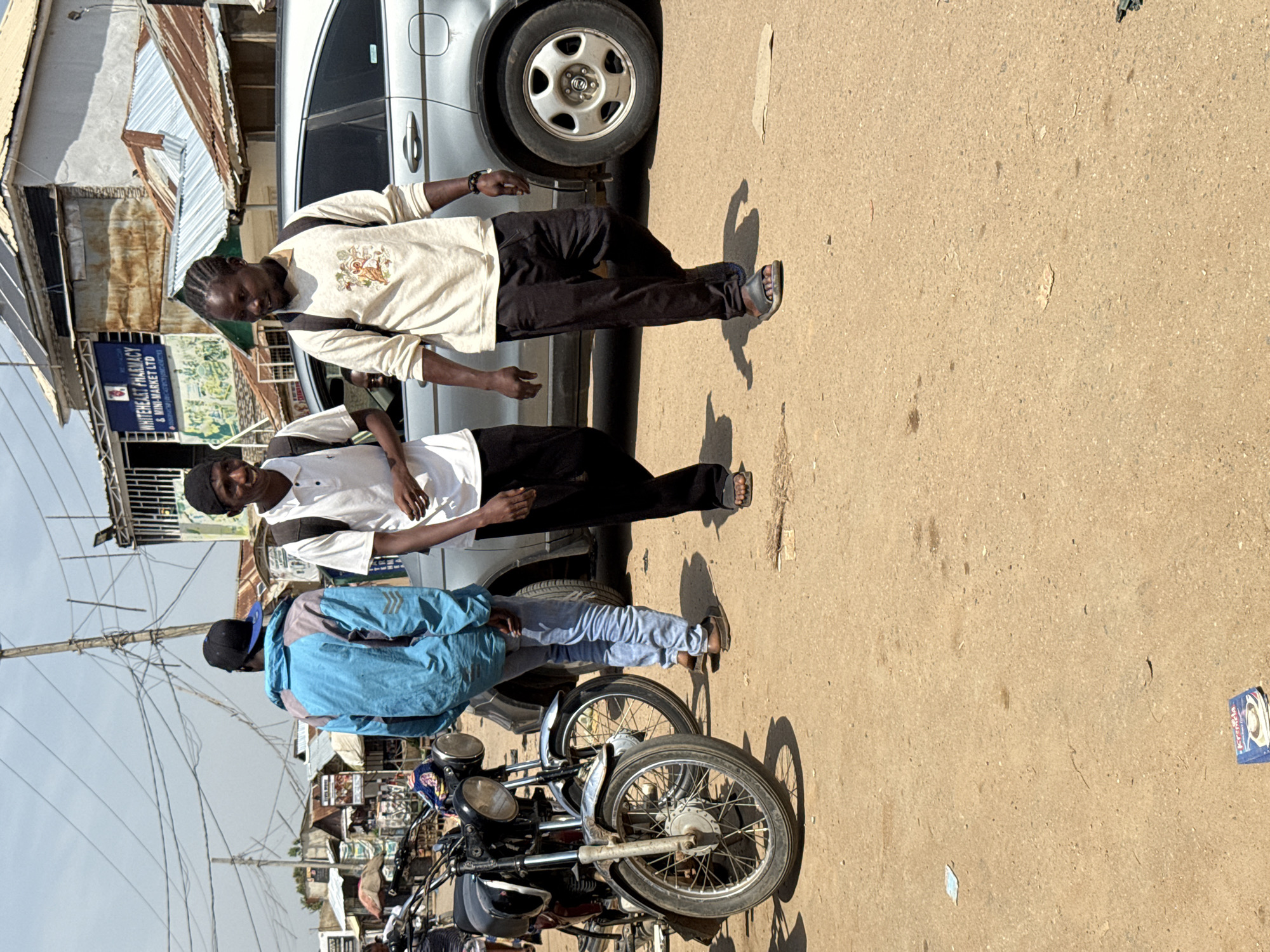 Two young men are walking on a dirt road in what appears to be a busy market area in Abuja, Nigeria. The man on the left is wearing a blue and grey jacket and a black baseball cap, walking towards the left of the frame. The man on the right has braided hair, wears a light-colored long-sleeved shirt with a graphic design and black pants, and is walking towards the right of the frame. Behind them, a silver SUV is parked, and to the left, a motorcycle is visible. The background shows several market stalls with corrugated metal roofs, and overhead power lines crisscross the sky. The ground is dusty and appears to be unpaved.