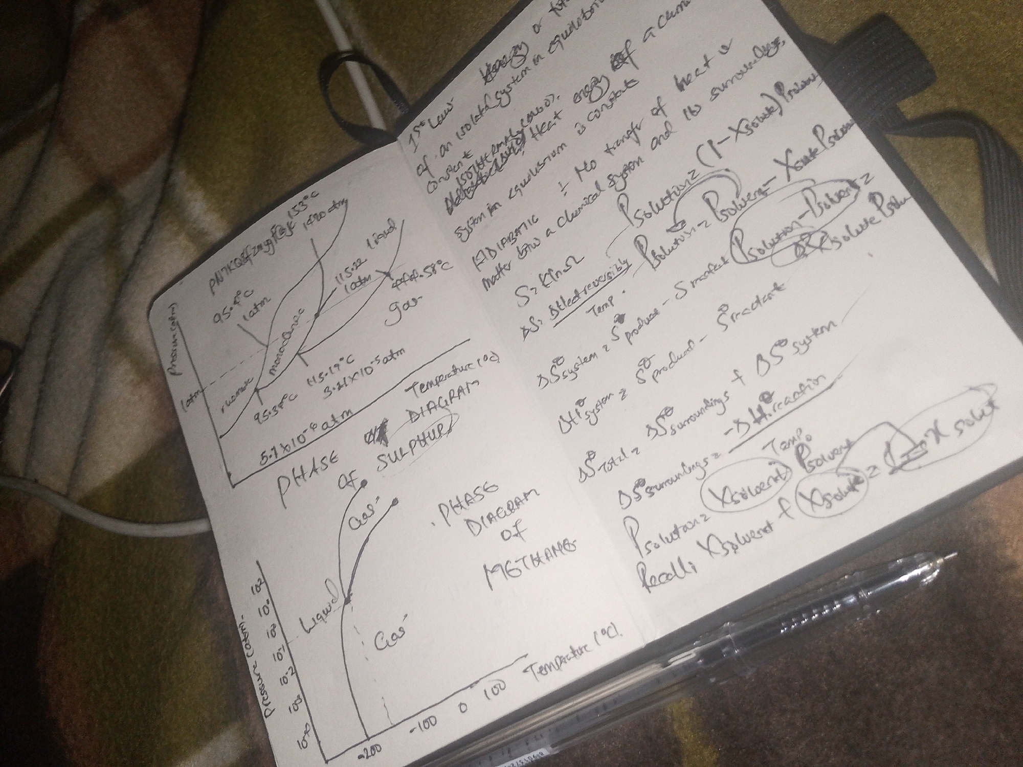 The image shows a close-up of an open notebook with handwritten notes and diagrams. The notebook is resting on a textured, patterned surface, possibly a blanket or upholstery, in a dimly lit environment. A black pen lies across the lower portion of the notebook, and a dark object, possibly a notebook cover or strap, is visible in the upper right corner. The handwritten content appears to be scientific notes, including equations, graphs, and chemical formulas. The notes are written in black ink on the white pages of the notebook. There are no people or recognizable landmarks visible in the image. The setting is a still life focused on the notebook and its contents.