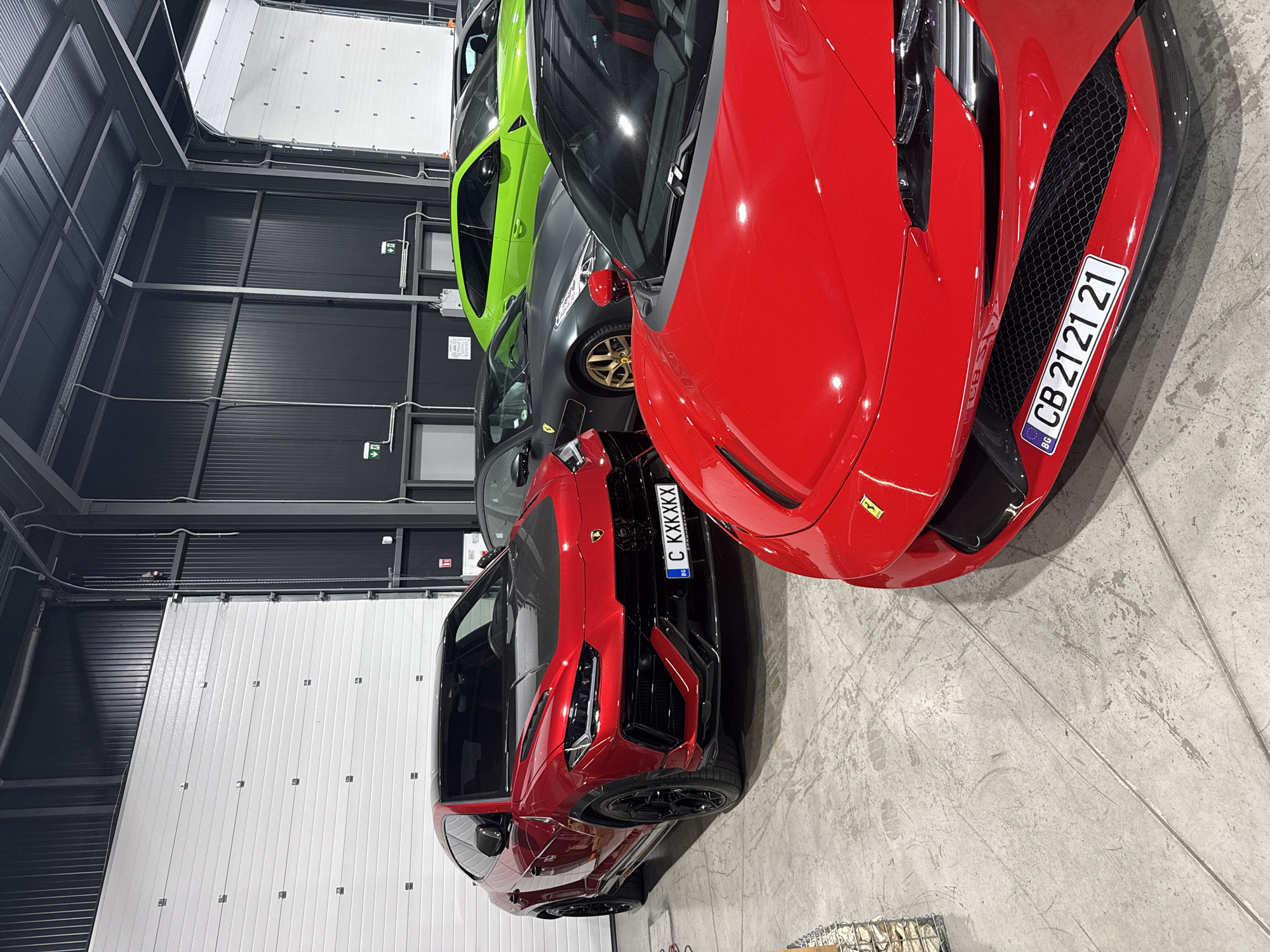 The image depicts an indoor garage or showroom in Plovdiv, Bulgaria, showcasing multiple high-performance automobiles. The primary subjects are four luxury vehicles, arranged on a light gray concrete floor marked with wear and expansion joints.
In the immediate foreground, a red Ferrari, likely a modern model such as an SF90 Stradale, is partially visible, primarily its front bumper and hood. It features a black honeycomb grille, carbon fiber lower lip, and a Bulgarian license plate "BG CB 21 21 21". The "CB" district code indicates Plovdiv.
To the left and slightly behind the foreground Ferrari, a red Lamborghini Urus SUV is prominently displayed, facing towards the right foreground. This vehicle features a dark carbon fiber hood, black trim on the front bumper and wheel arches, and black multi-spoke wheels. Its Bulgarian license plate reads "BG C KXXXXX".
Behind the foreground Ferrari and to the right, a dark gray Ferrari convertible, possibly a Portofino or Roma, is visible, distinguished by its gold-colored multi-spoke wheels. Further in the background, a bright lime green Lamborghini Urus is parked, partially obscured.
The environment consists of dark gray/black corrugated metal panel walls and large white sectional garage doors on the left and right. Exposed silver-colored conduits, pipes, and structural beams are visible along the walls and ceiling, indicative of an industrial or warehouse setting. Green "exit" signs with downward arrows are affixed to the dark gray walls. No people are present in the scene.